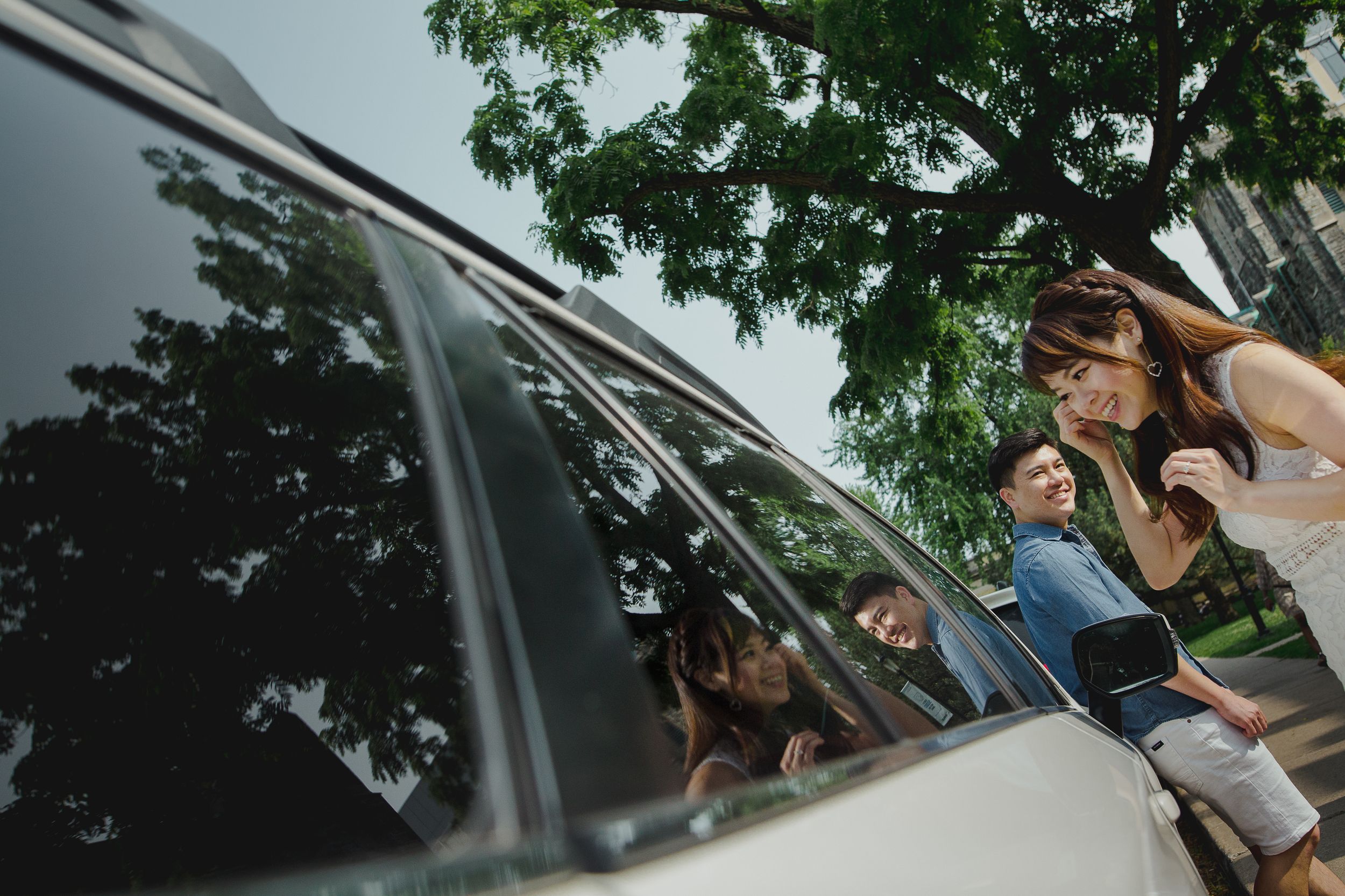 A person in a white shirt leans through a car window on a sunny day with trees visible in the background.