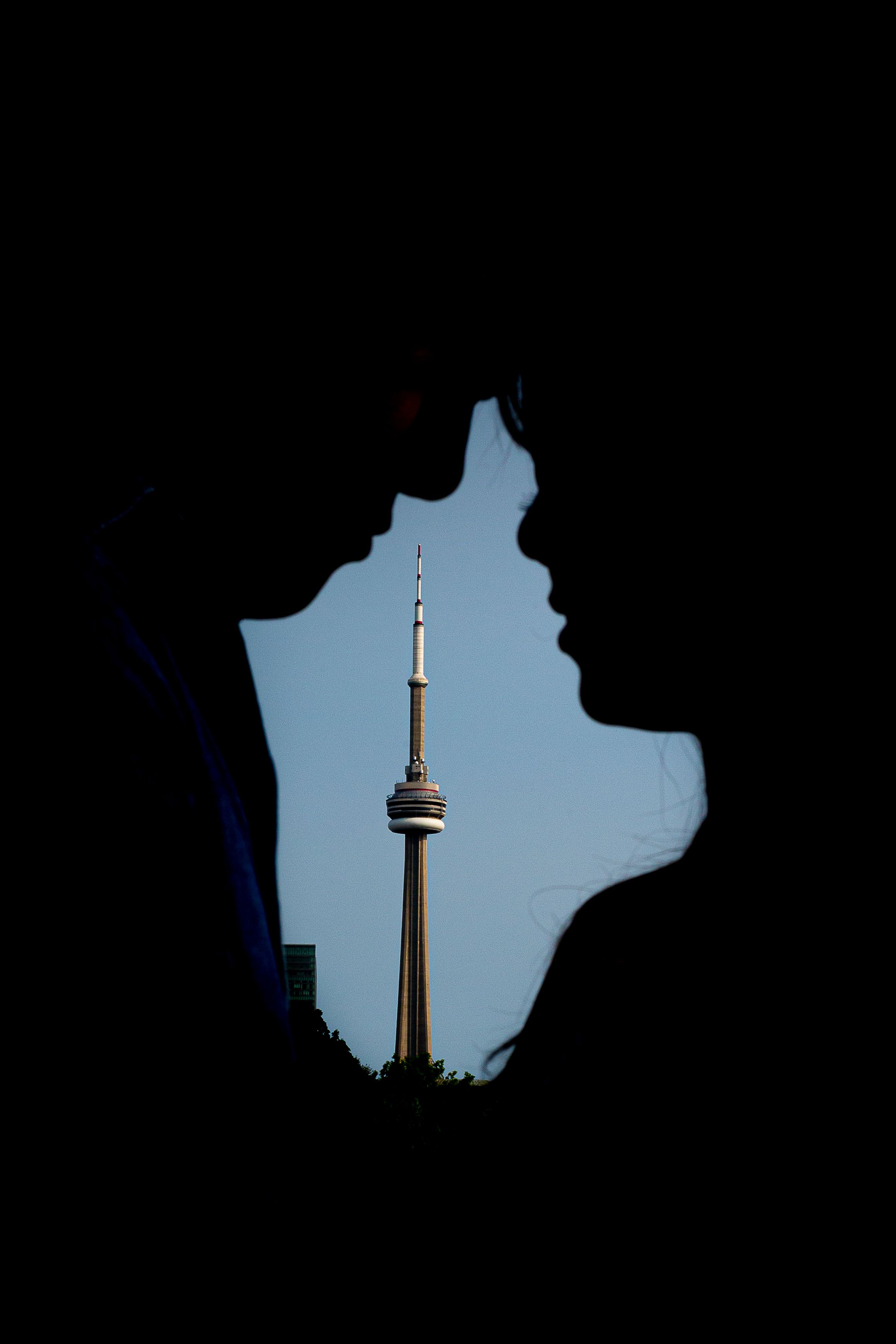 Silhouetted faces in profile create negative space revealing the CN Tower against a blue sky in Toronto.