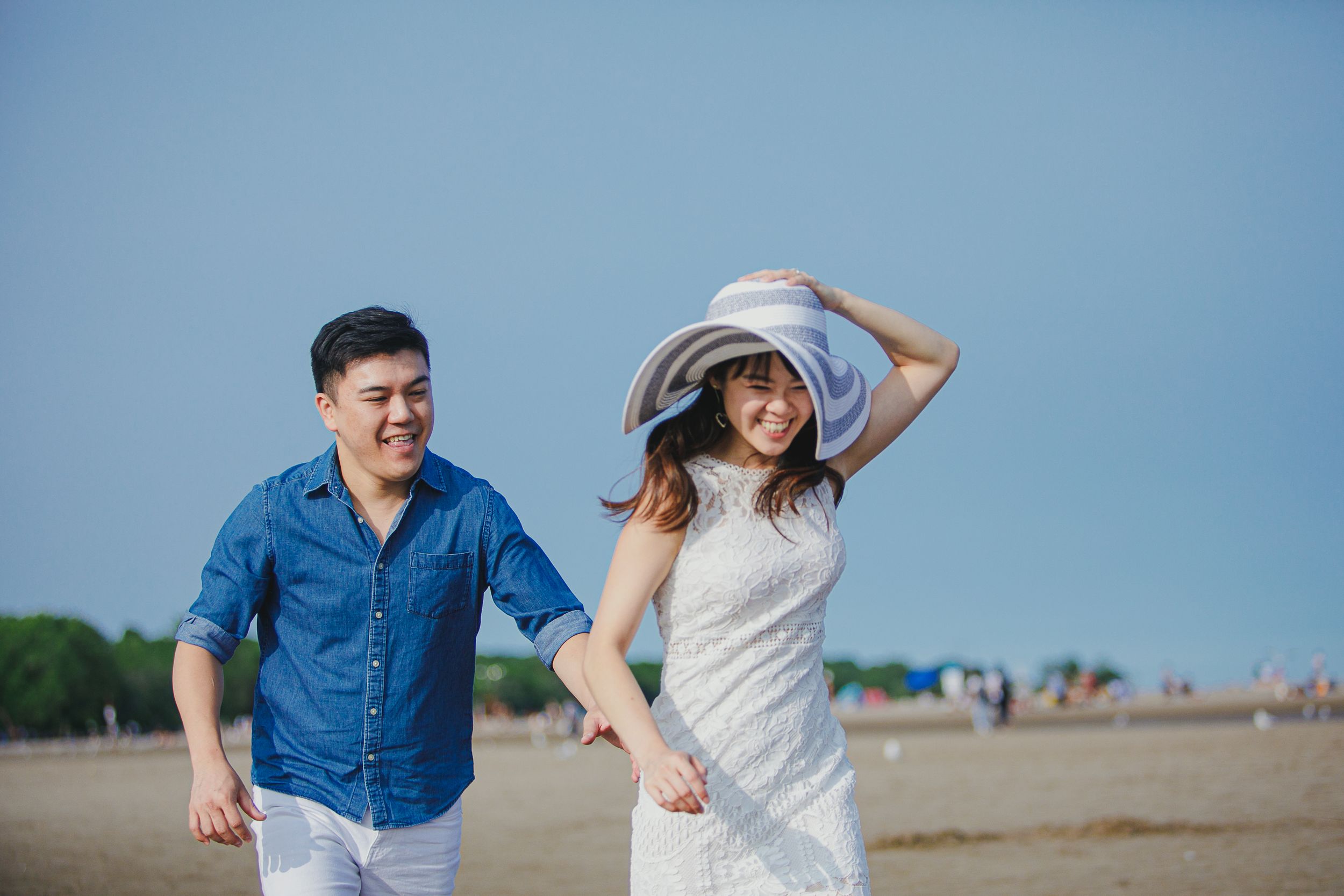 A couple in blue and white outfits walk joyfully along a sandy beach on a sunny day.