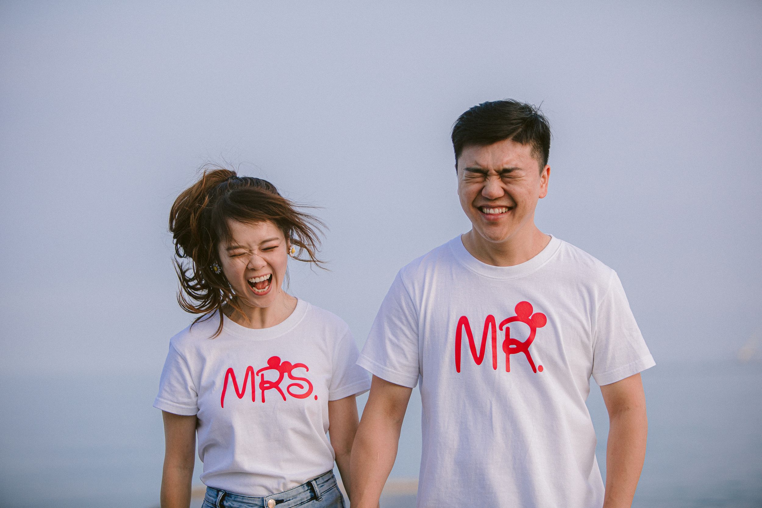 A couple in matching white t-shirts with 'Mr' and 'Mrs' text sits together by the ocean.