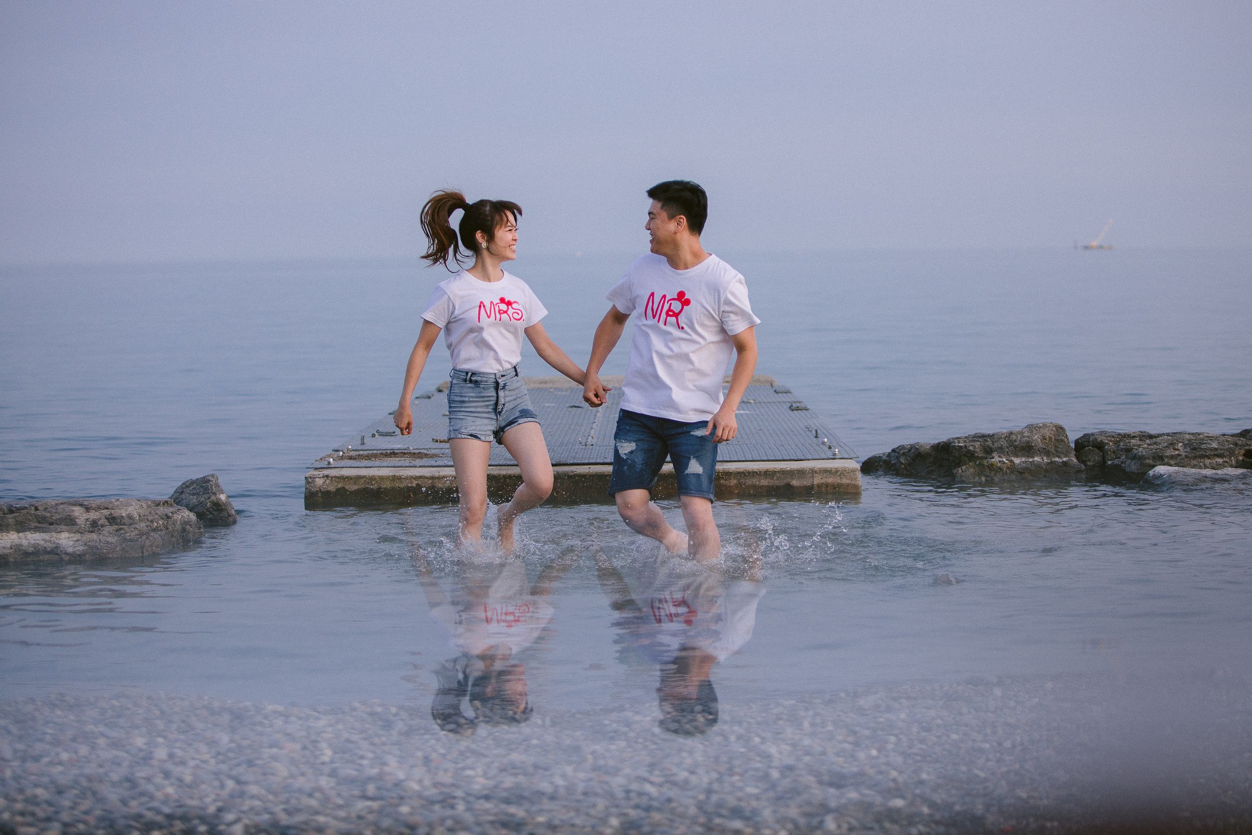 Two people sit casually on rocks by the waterfront wearing matching white shirts.