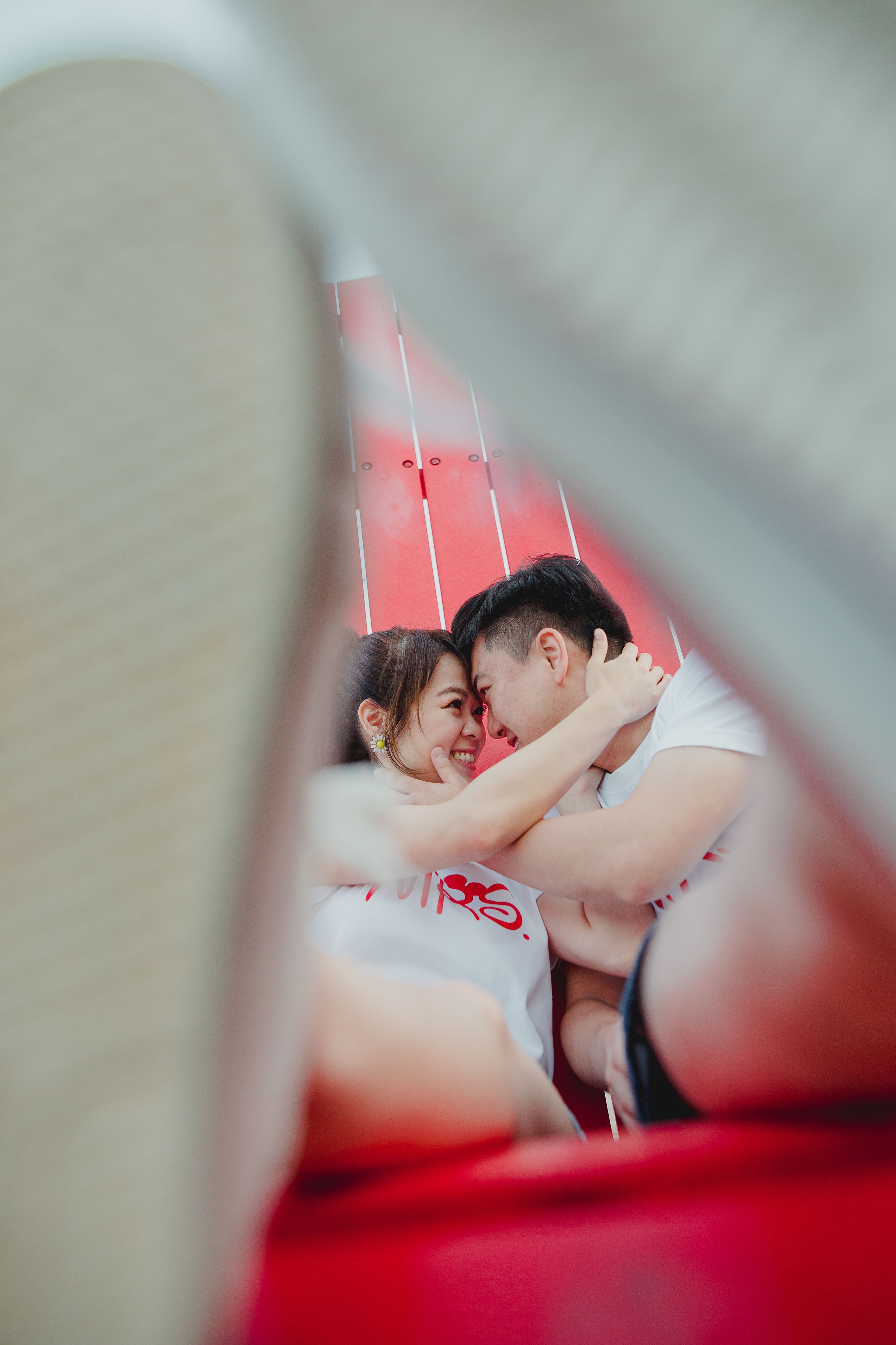 Two people recline together on a bright red wooden bench near the ocean.