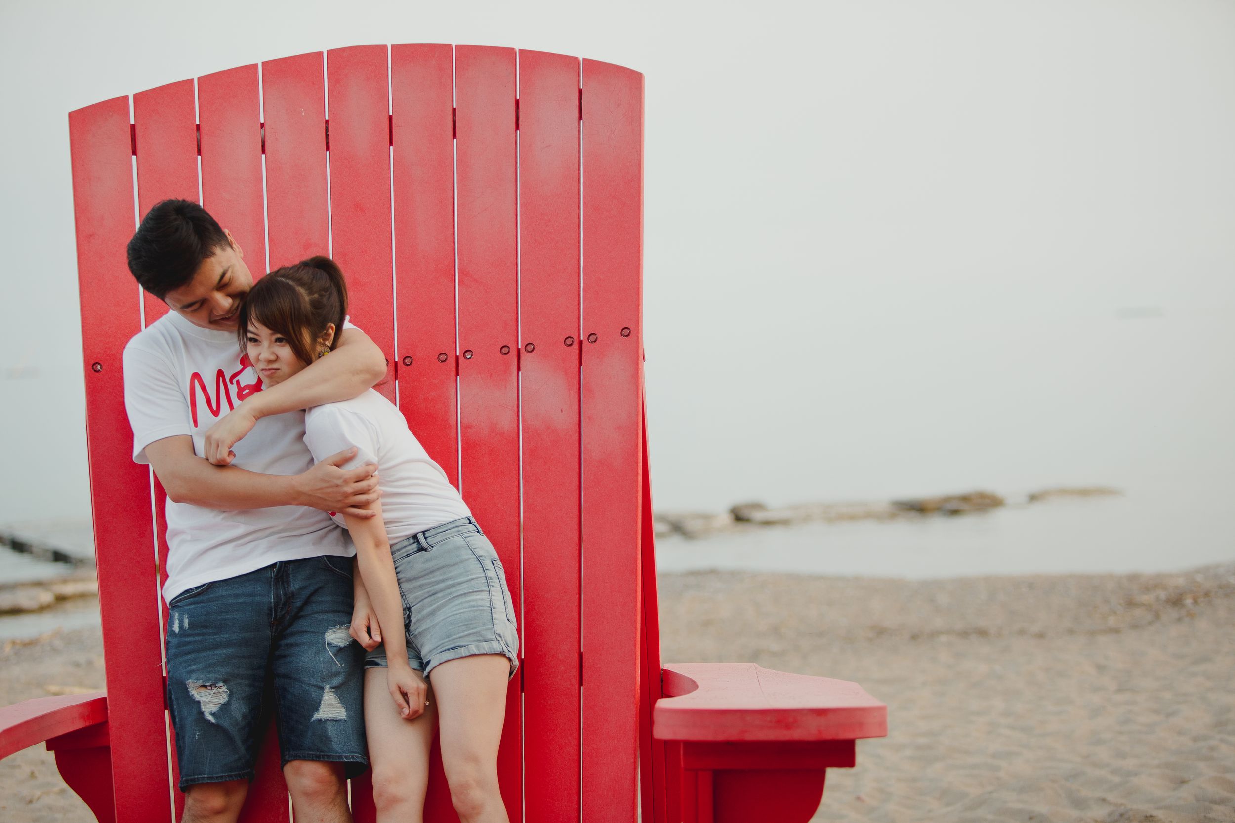 A couple embraces while sitting on a vibrant red wooden bench at the shoreline.