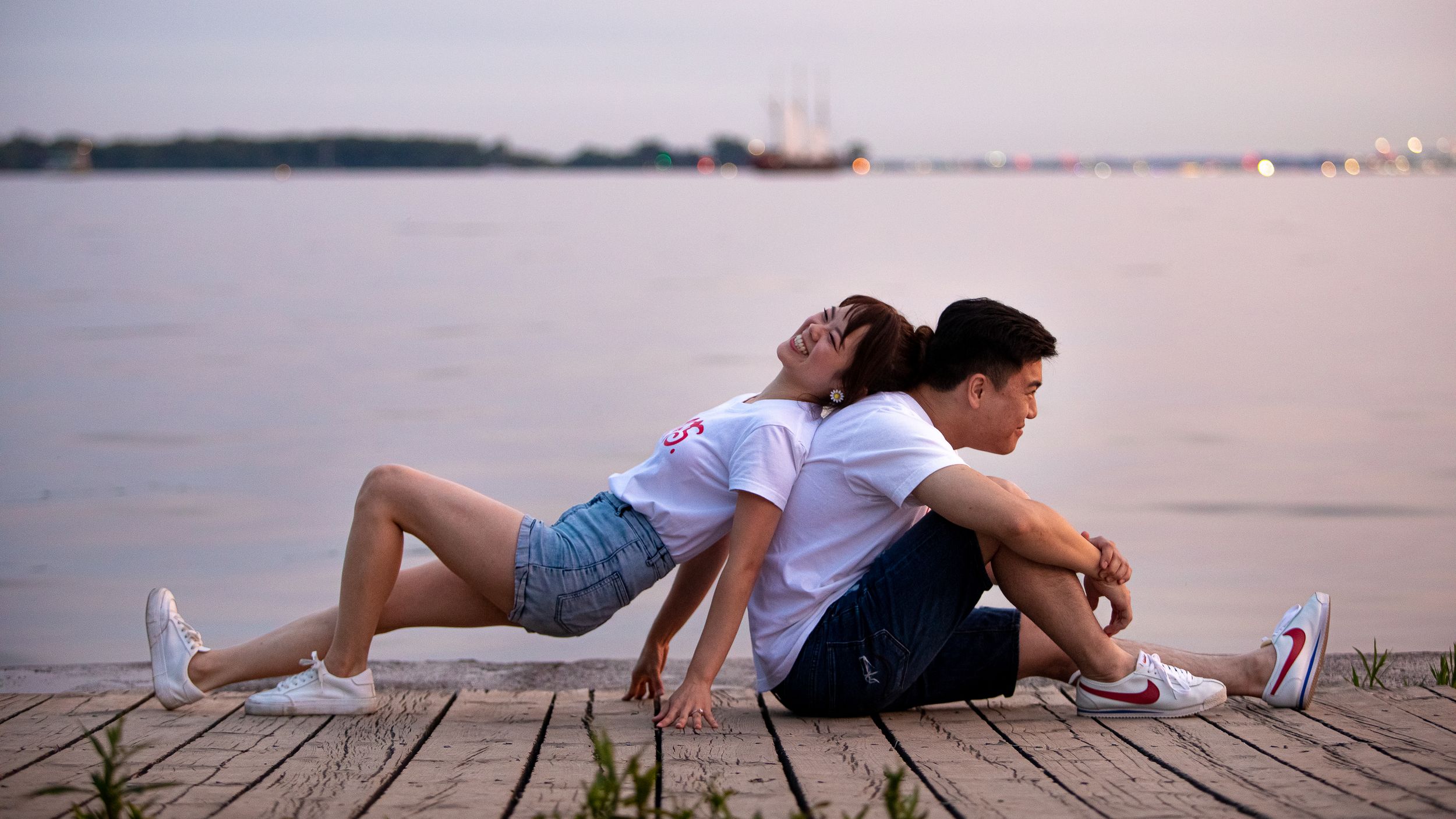 A romantic couple lies together on a wooden pier overlooking a peaceful lake at sunset.