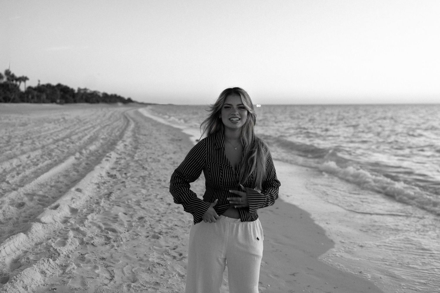 Black and white photo of someone smiling while standing on a sandy beach at sunset.