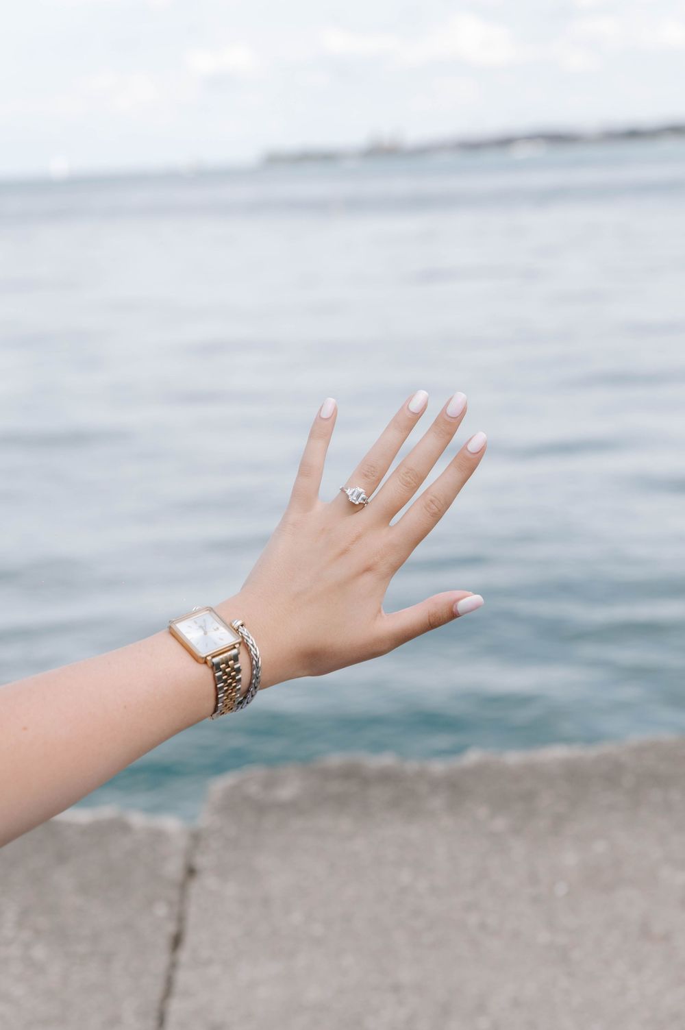 A hand with pale pink manicured nails and an engagement ring reaches out over ocean waters, wearing a gold watch.