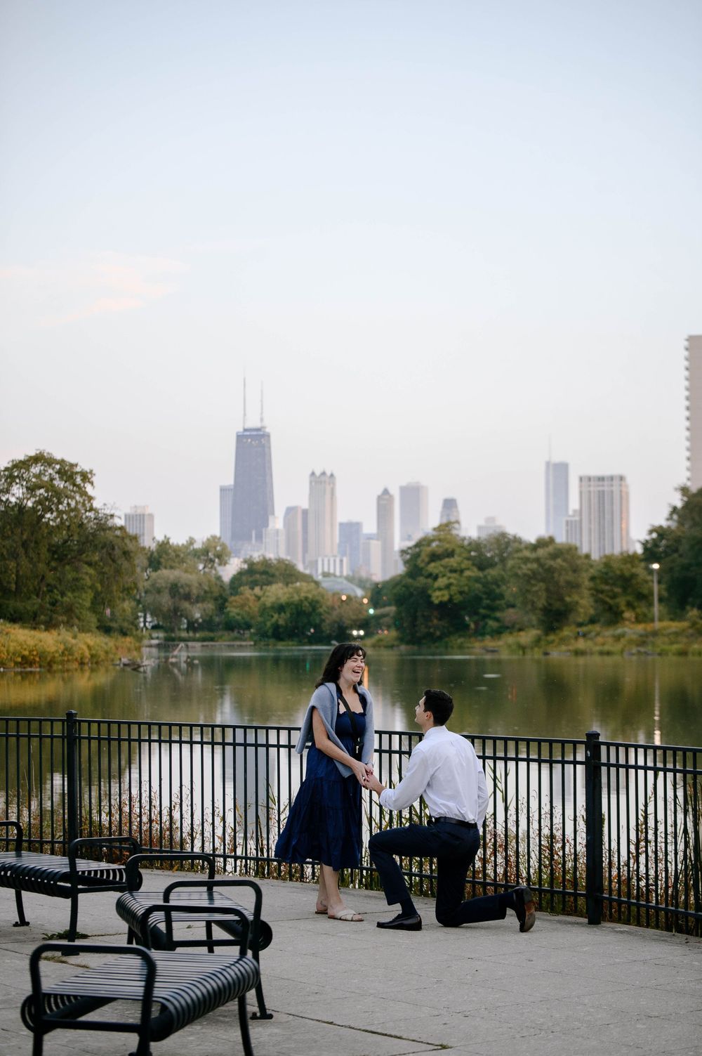A couple sits on benches overlooking a city skyline and lake in a romantic park setting during autumn.