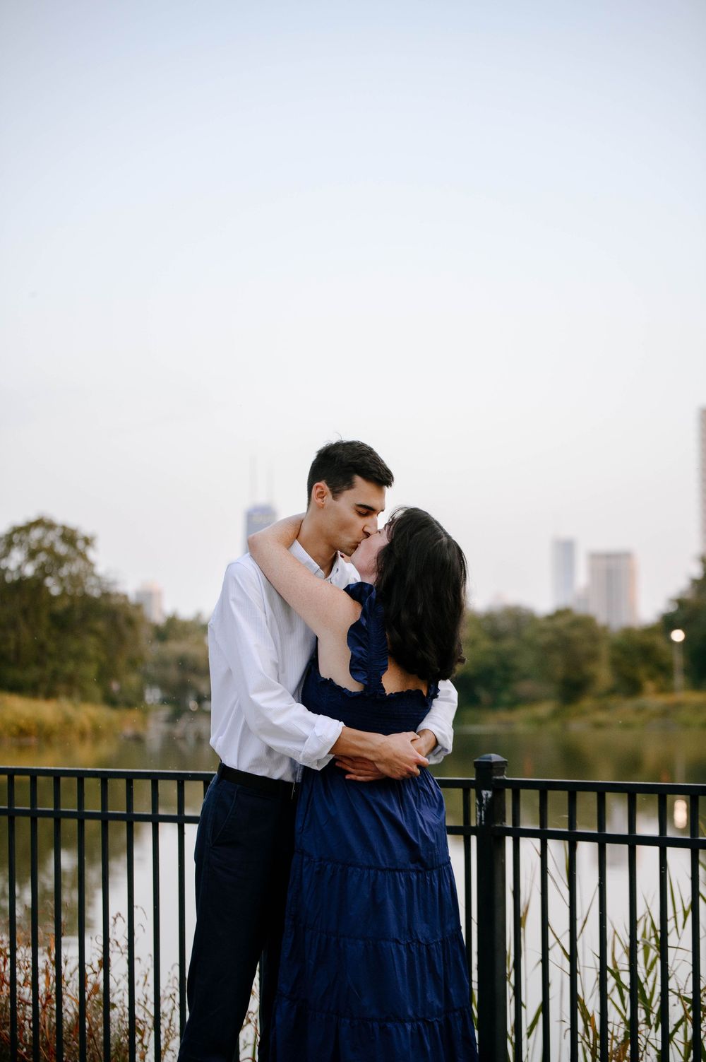 A romantic couple shares intimate moments by a lakeside railing during a sunset photoshoot in an urban park setting.