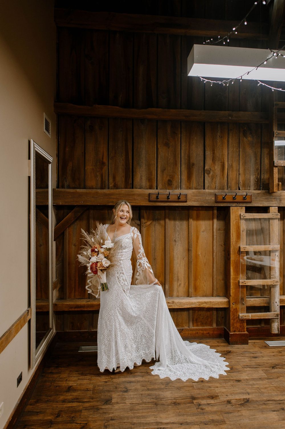 A bride in a long-sleeved lace wedding gown holds a bouquet while standing against a rustic wooden wall.