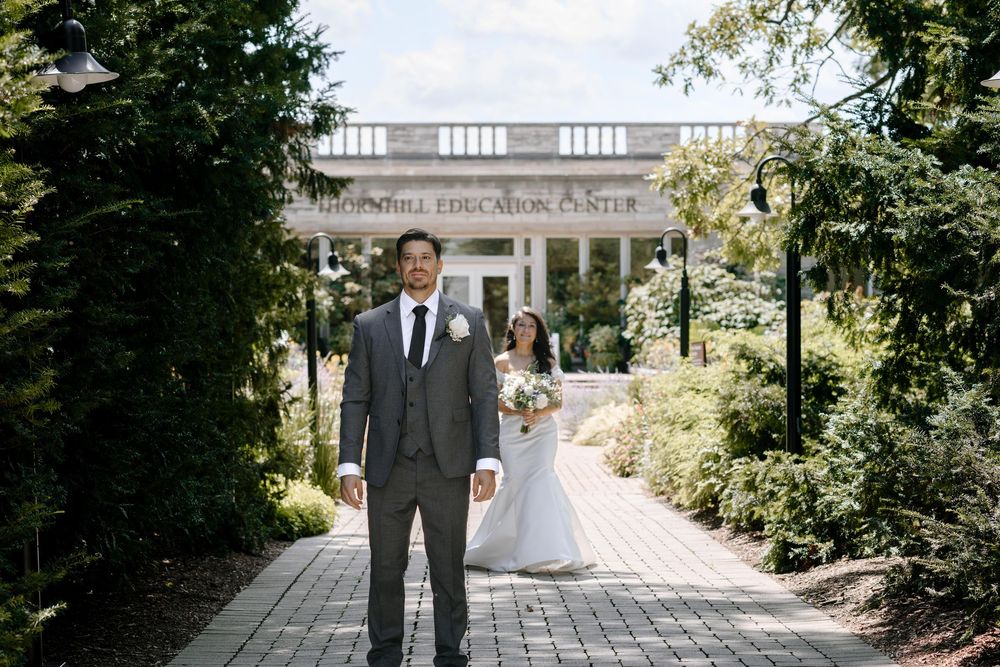Couple in formal attire walking down a garden path with lush greenery and stone architecture in the background.
