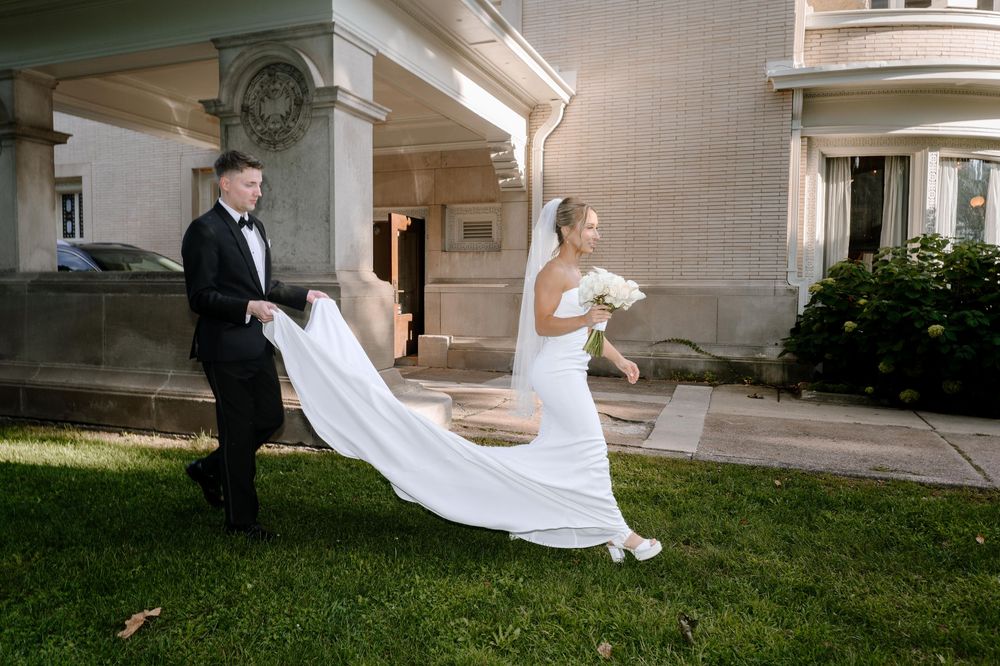 A flowing white wedding dress train trails behind a bride on a grassy lawn outside an elegant building.