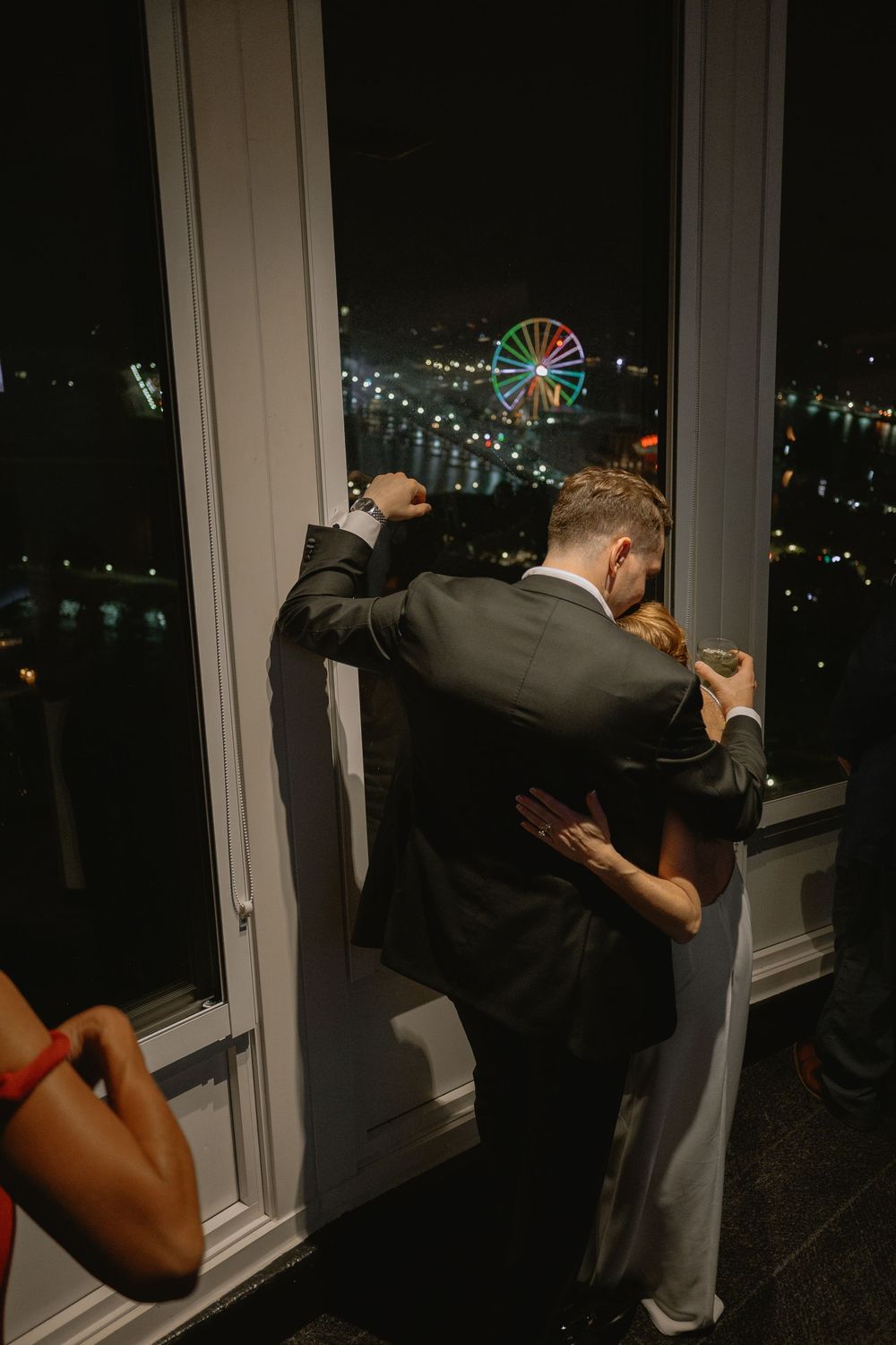 Couple embracing by a window at night with a colorful illuminated Ferris wheel visible in the city skyline below.