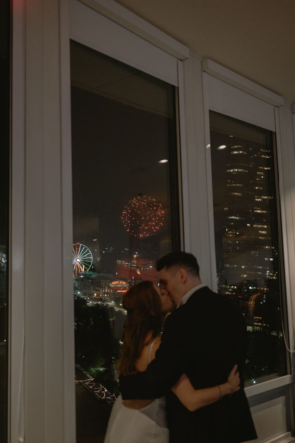Couple sharing a romantic kiss in front of city skyline with fireworks and Ferris wheel at night.