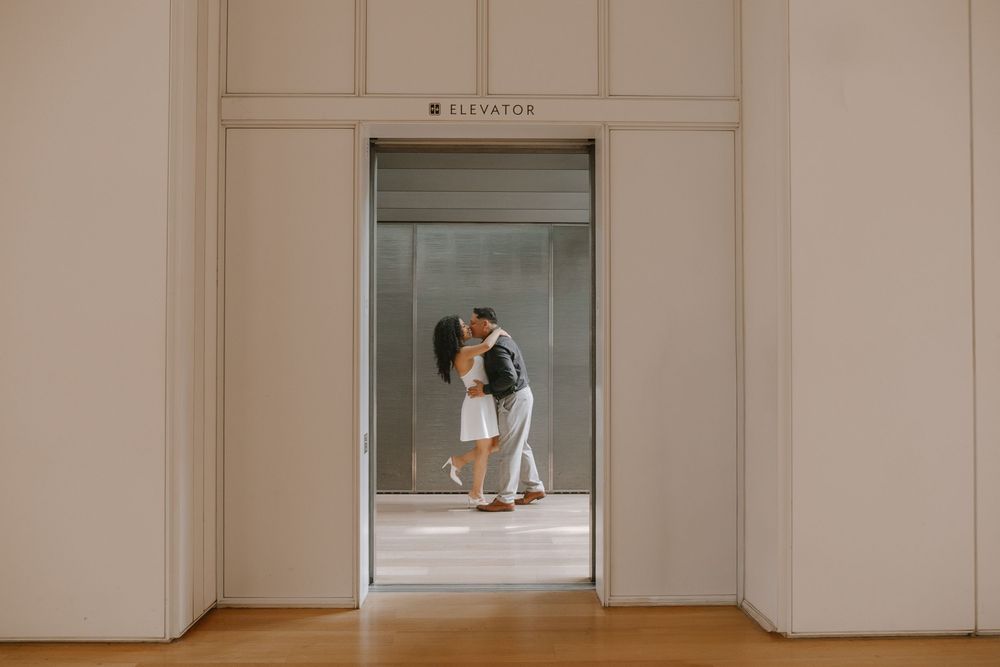 Couple sharing a romantic kiss in front of an elevator in an elegant building with wooden floors.