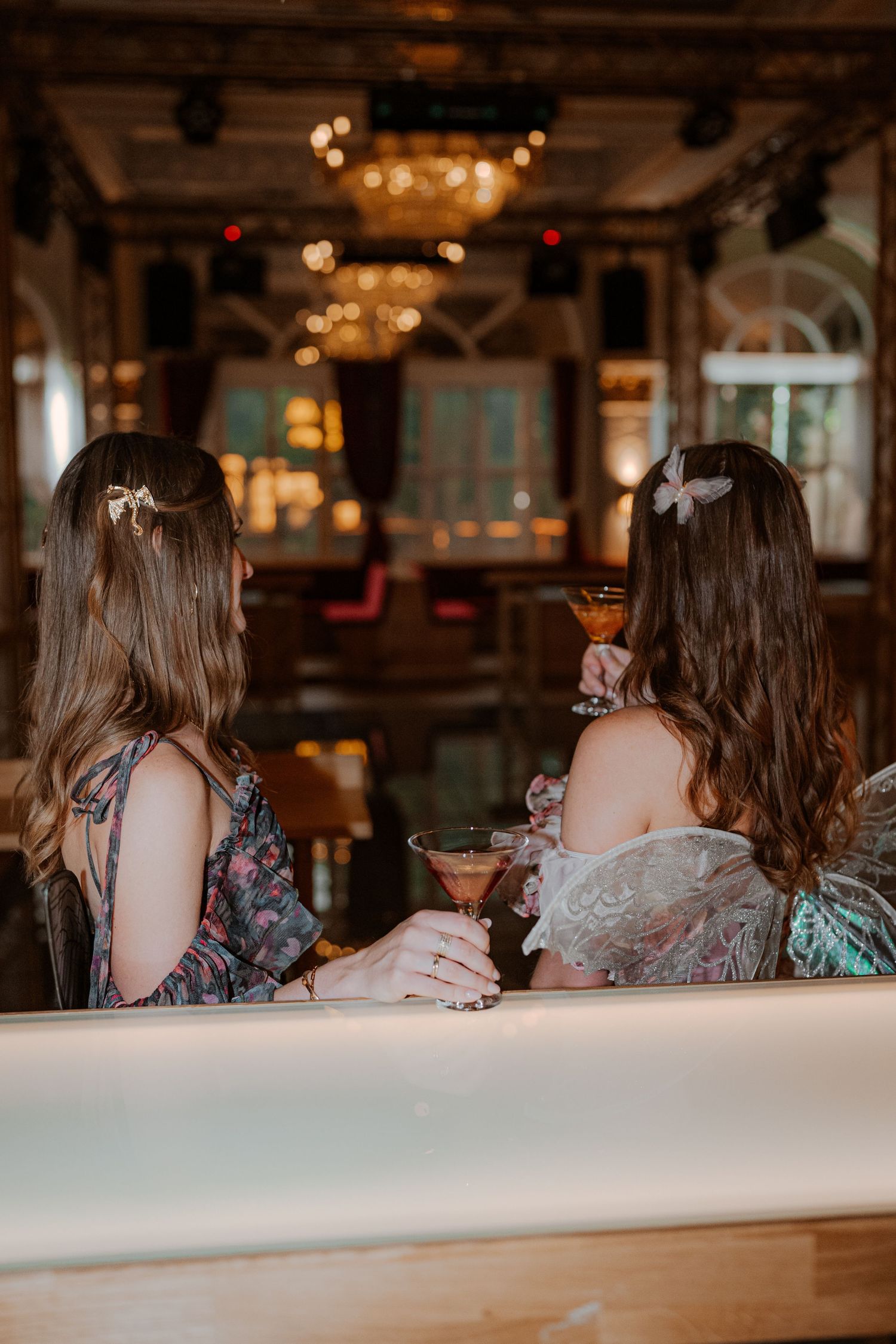 Two women with long brown hair sit at a bar counter holding cocktail glasses in an elegantly lit establishment.