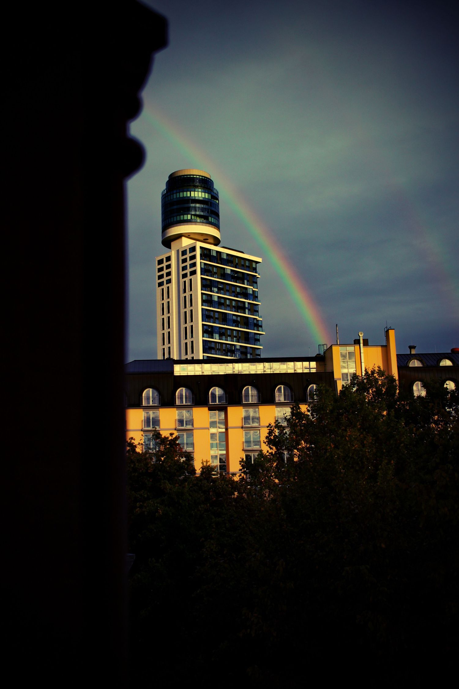 A rainbow arches over a modern office building complex against a dramatic stormy sky at sunset.