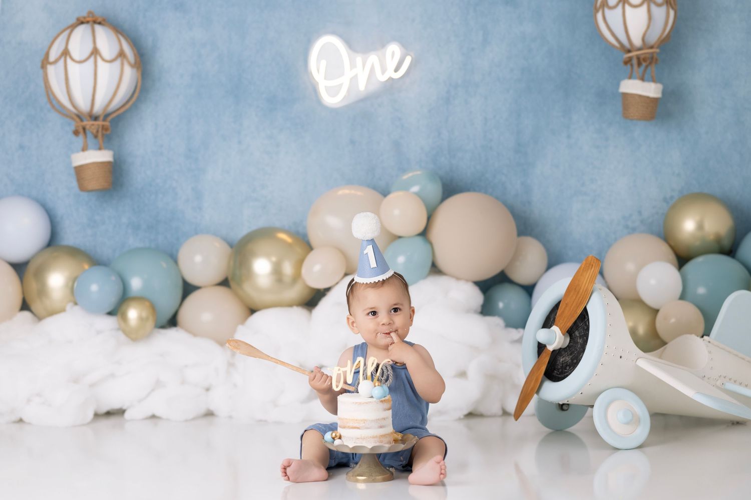 First birthday celebration setup with hot air balloon decor, pastel balloons and white clouds against blue backdrop.