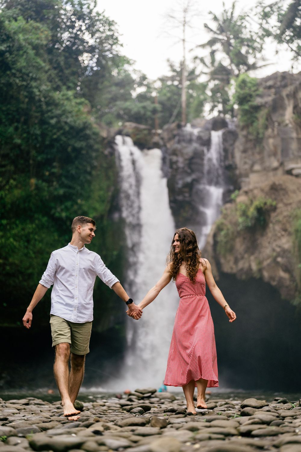 Couple playfully walking on rocky terrain with waterfall backdrop.