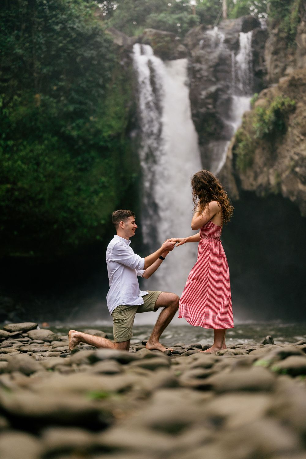 A romantic proposal moment unfolds by a majestic waterfall with a couple sitting on rocky terrain.