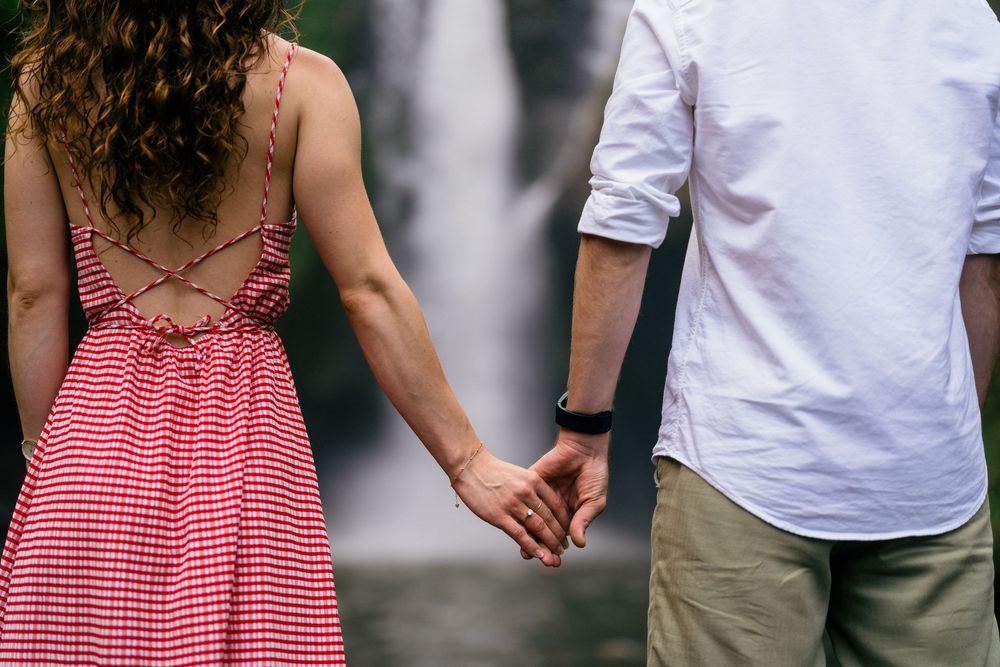 Two people in pink and white outfits stand together against a scenic waterfall backdrop.