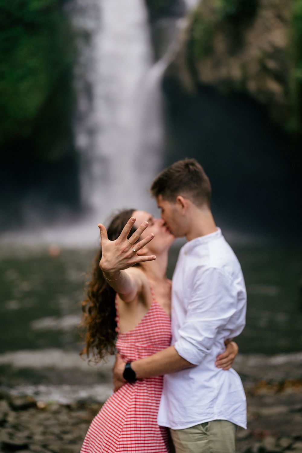 A tender embrace captured near a stunning waterfall with rocks in the foreground.