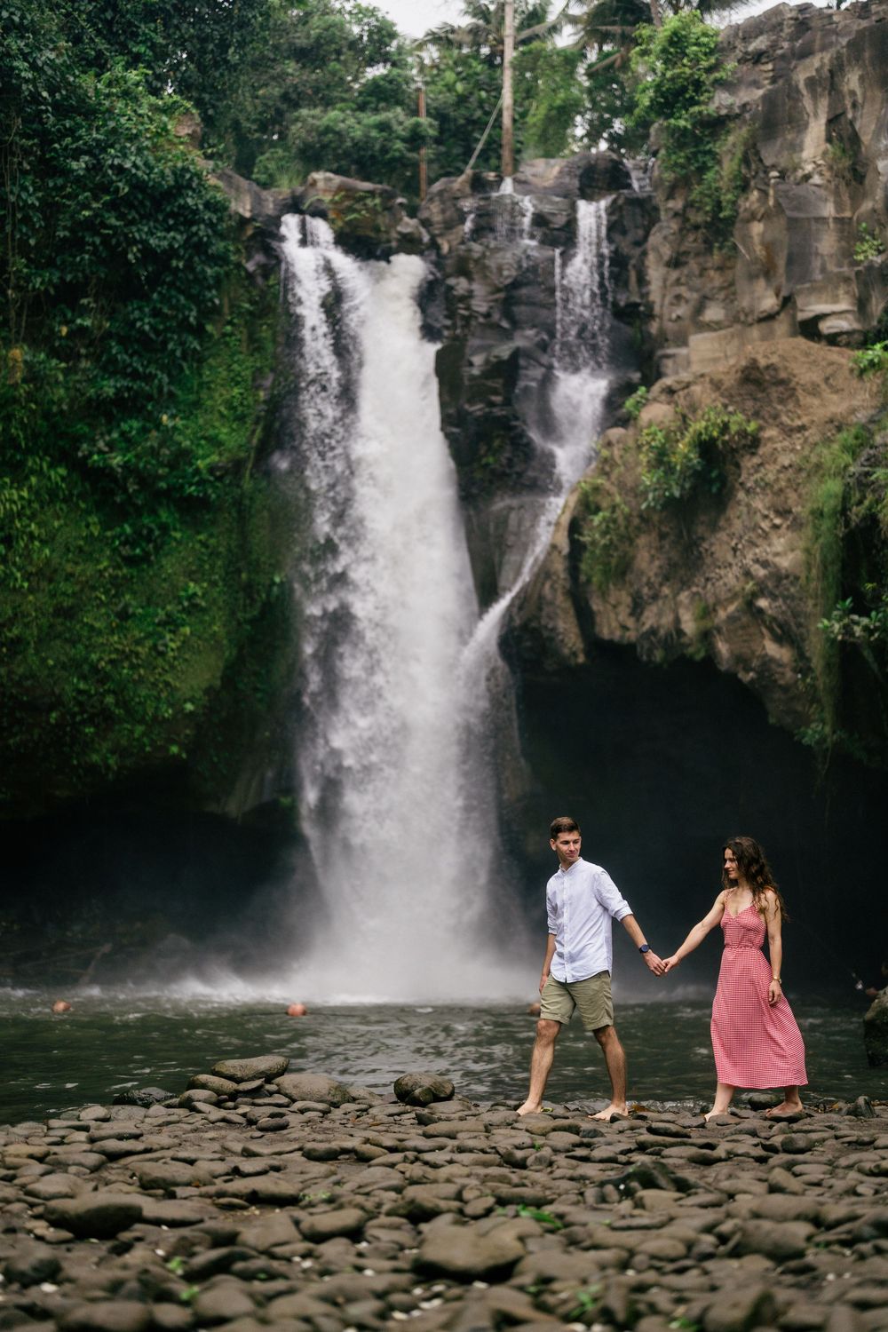 Two people walk hand in hand across rocky terrain with a dramatic waterfall in the background.