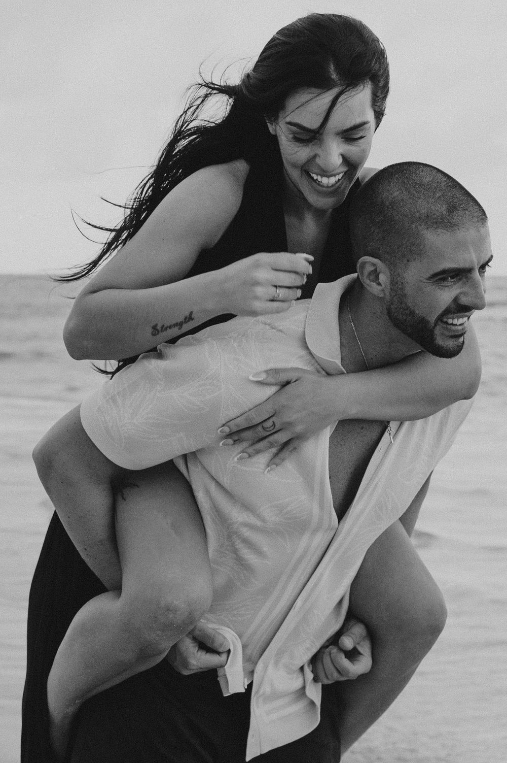 A joyful couple in black and white share a playful piggyback moment on the beach as the wind blows through their hair.