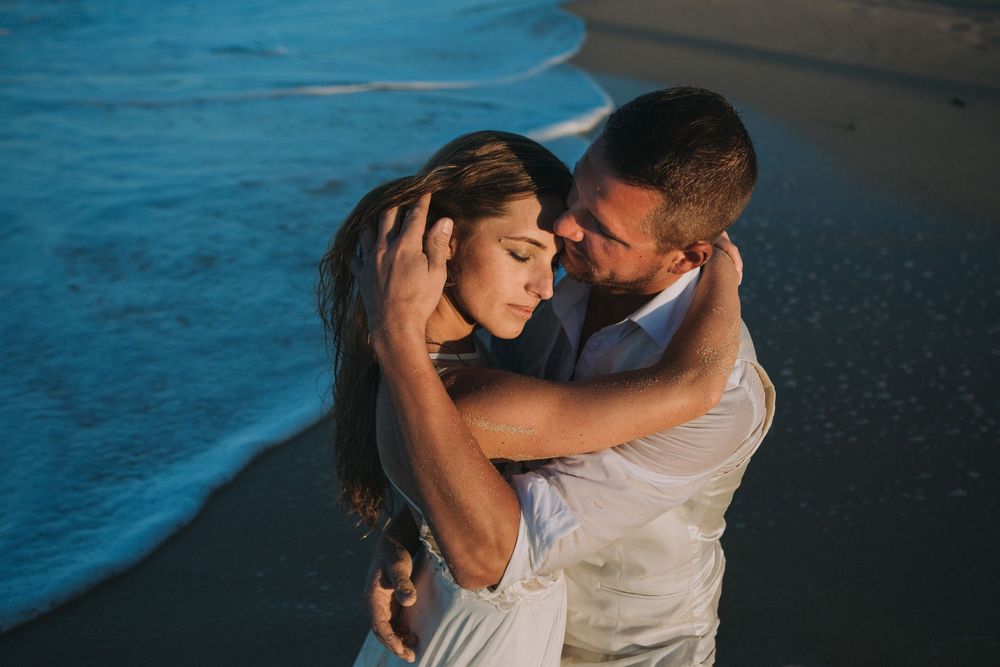 A couple shares a romantic moment along a beach shore at sunset with soft turquoise water in the background.