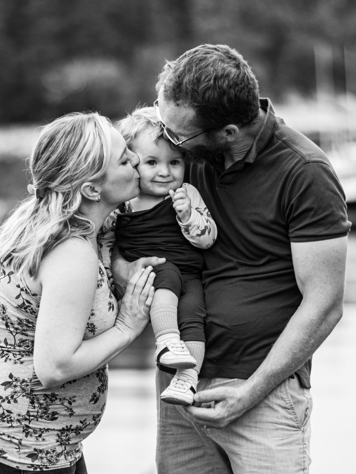 Black and white photo of parents kissing their happy toddler on both cheeks outdoors. Photo by Xenophon Photography in Maine.