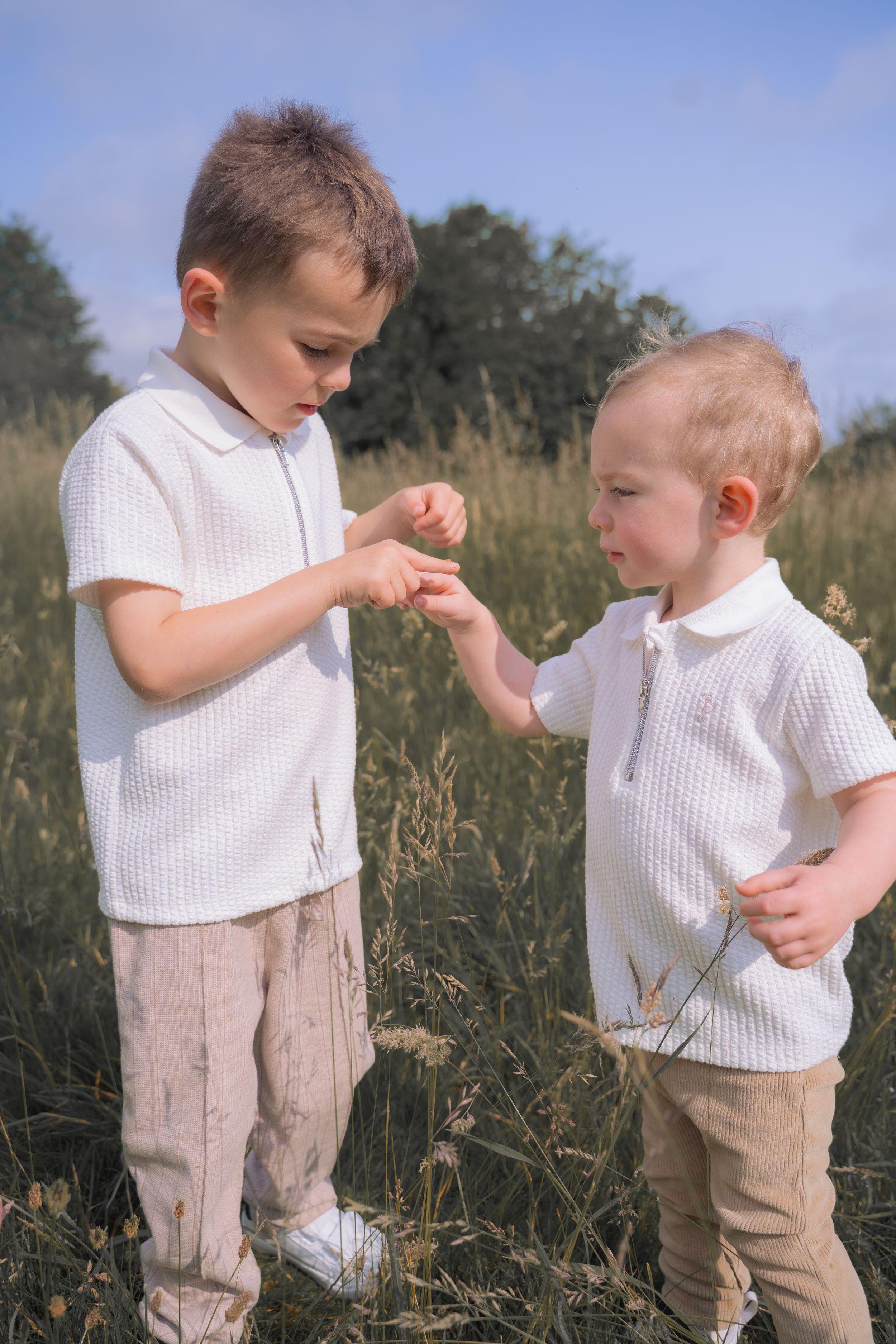 Two children in white shirts and khaki pants interact while standing in a grassy field on a sunny day.