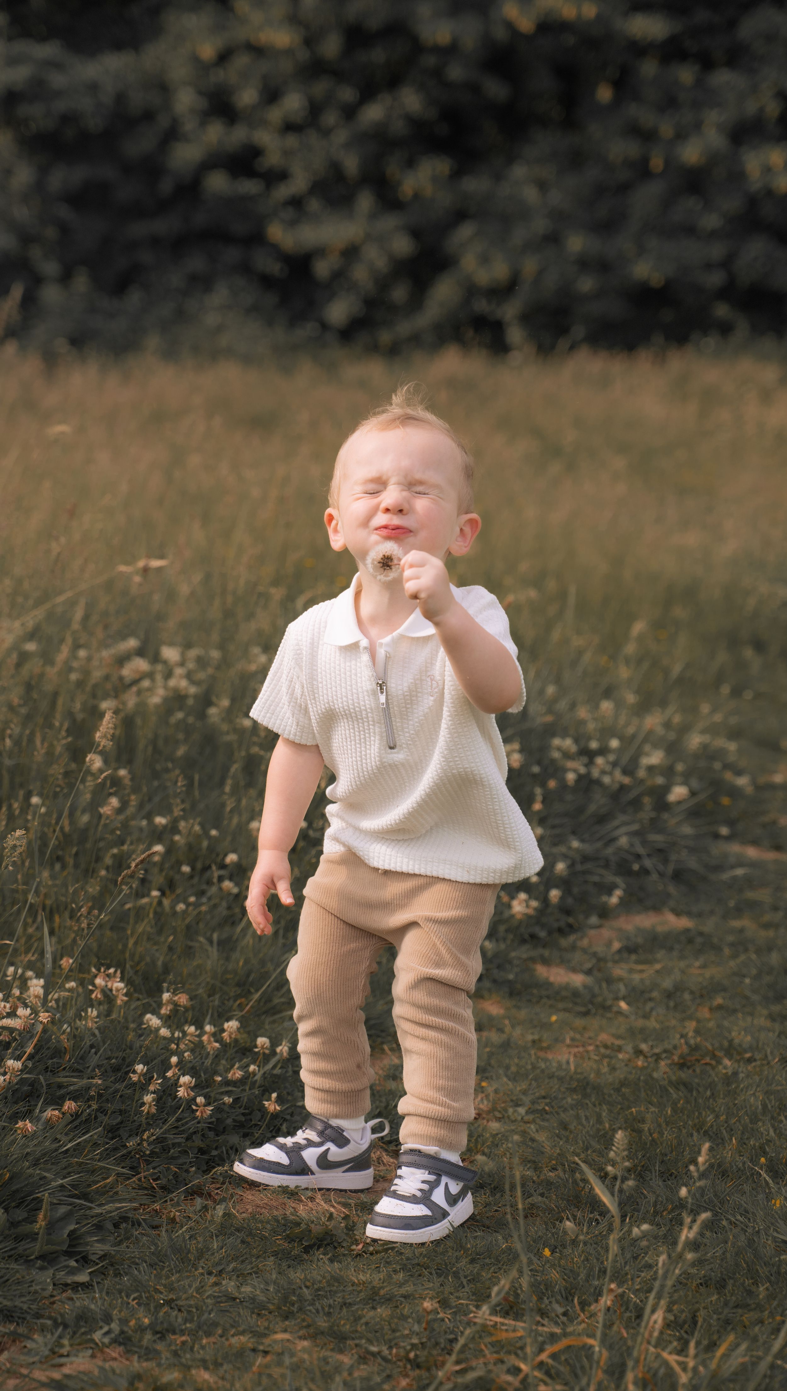 A toddler in a white shirt and beige pants laughs joyfully while playing outdoors in a wildflower field.