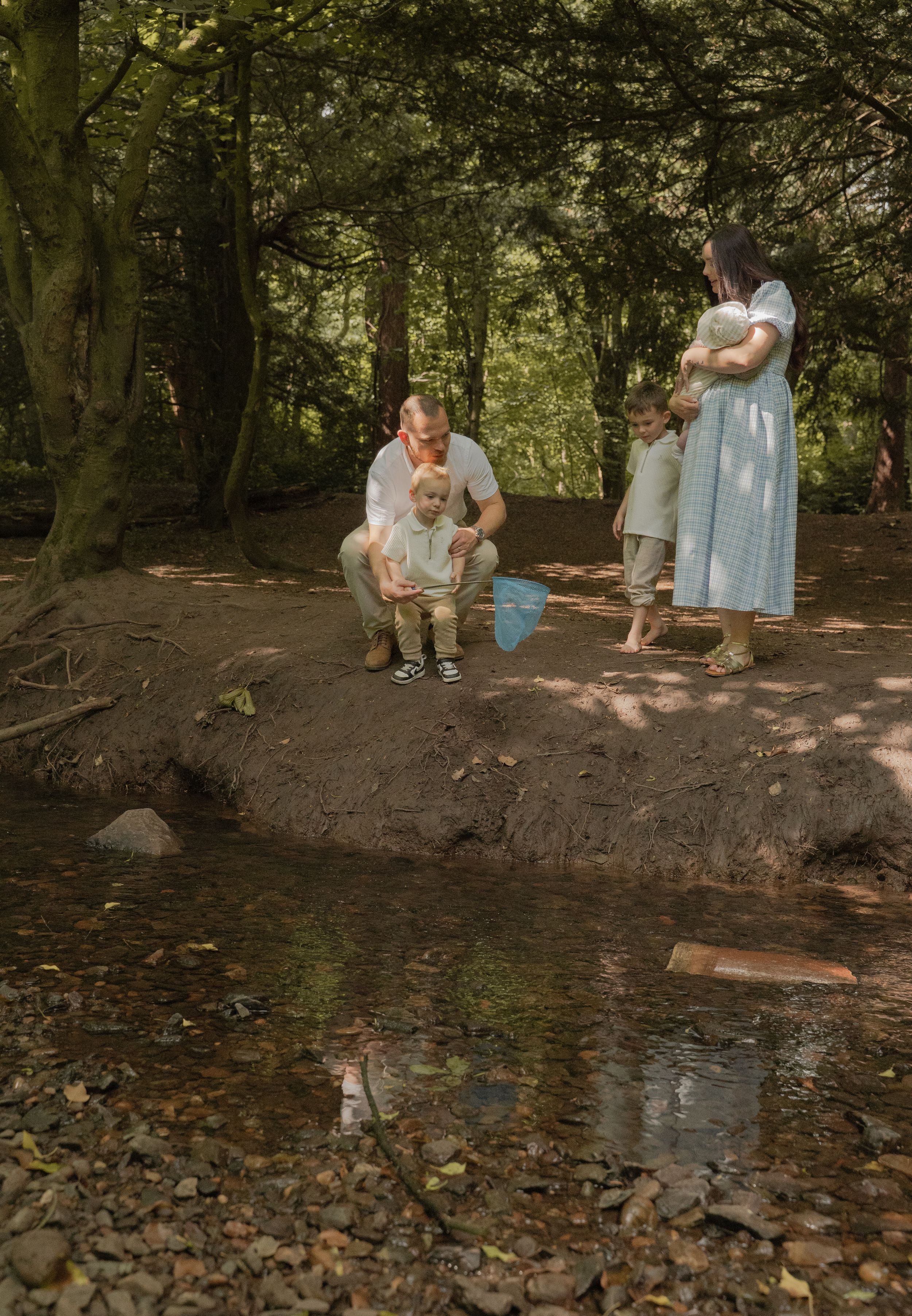 Two people sit by a peaceful stream in a wooded area during a sunny summer afternoon.