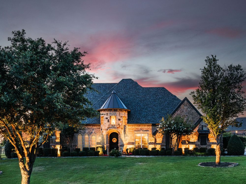 Luxurious estate home illuminated at dusk with landscaping lights and dramatic purple and pink sunset sky.