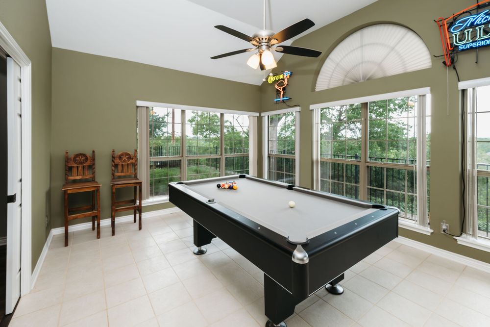 Bright game room with black pool table, tile floors, large windows, and ceiling fan in a spacious sunlit space.