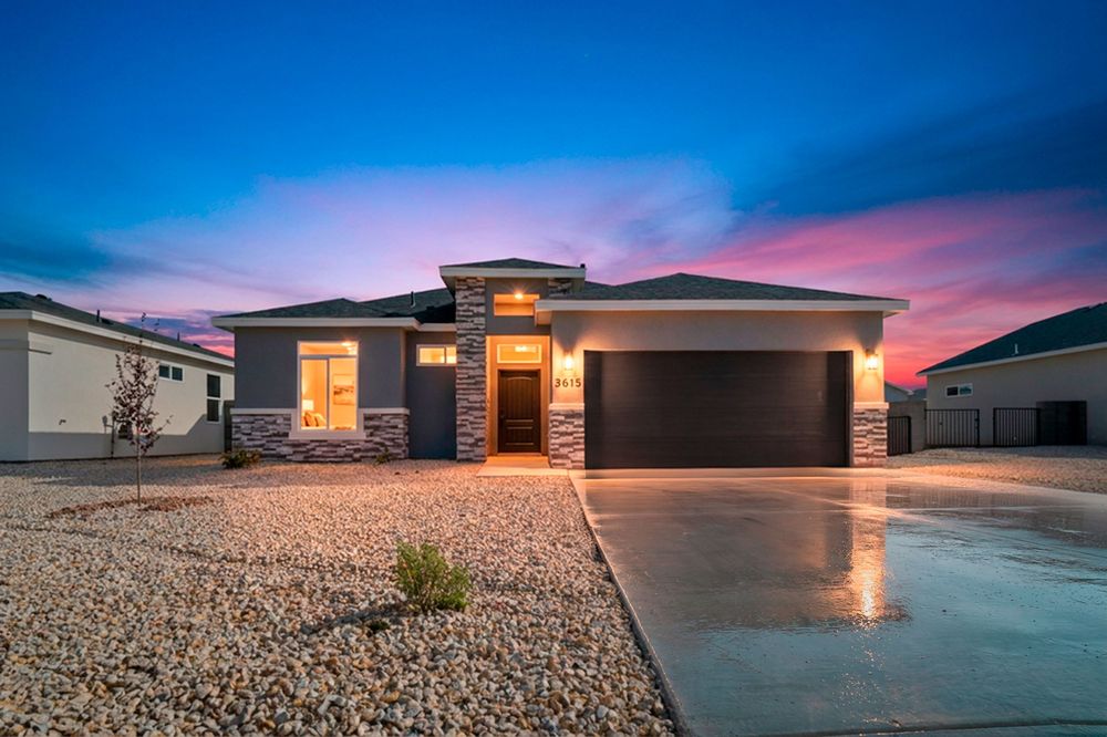 Modern single-story home with stone accents, lit entryway, and two-car garage under a vibrant sunset sky.