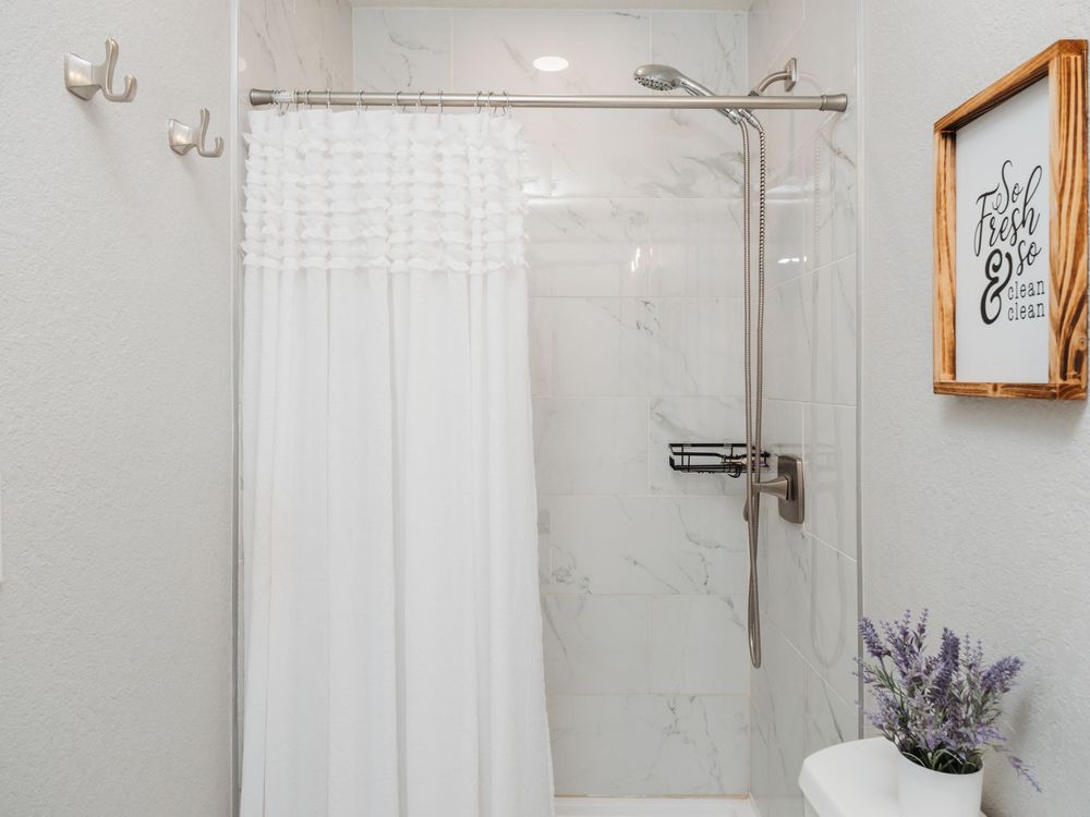 Modern white bathroom with marble shower, white curtain, brushed nickel fixtures, and lavender plant decor.