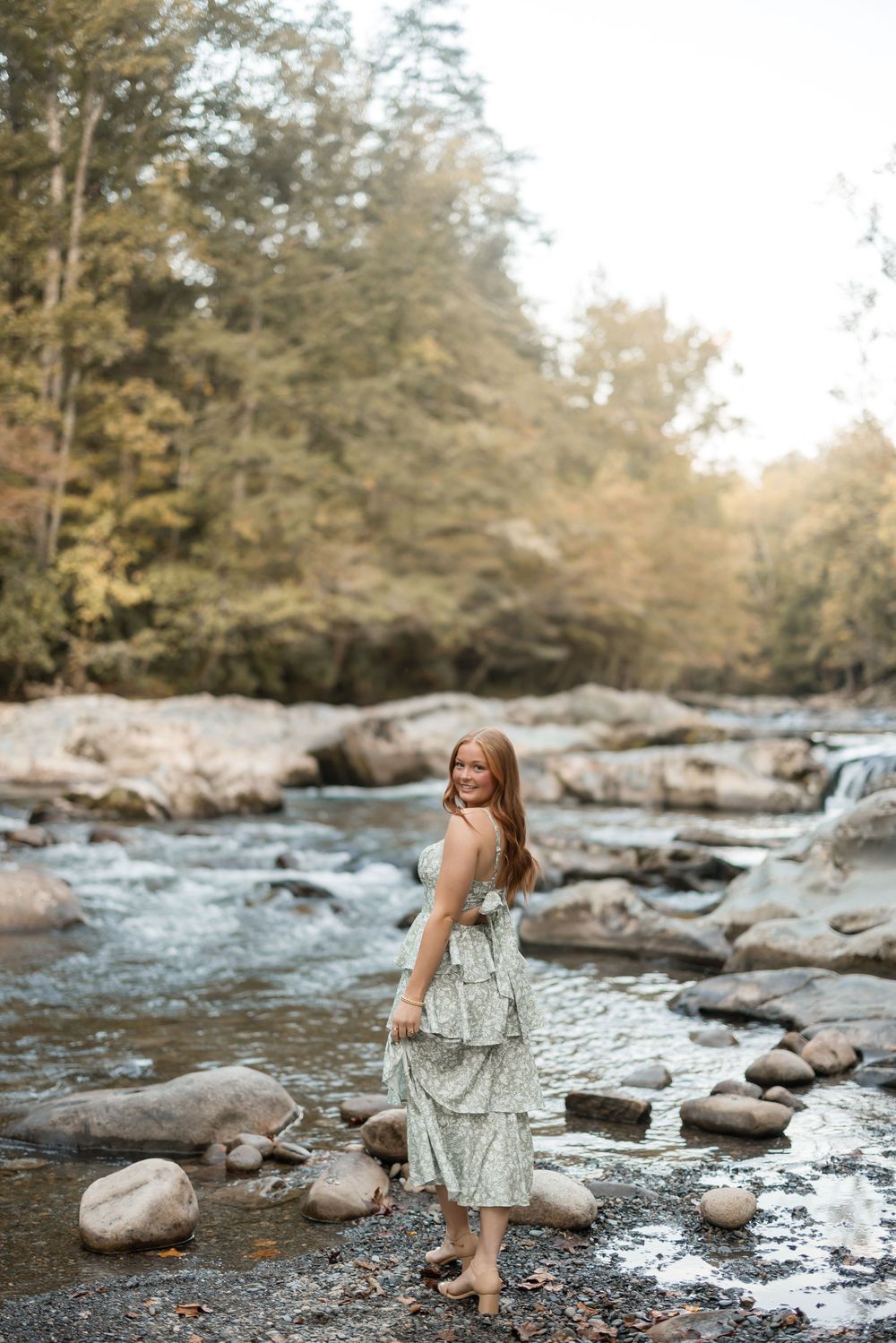 Someone in a floral dress explores a rocky riverbed with flowing water and large boulders in a forest setting.