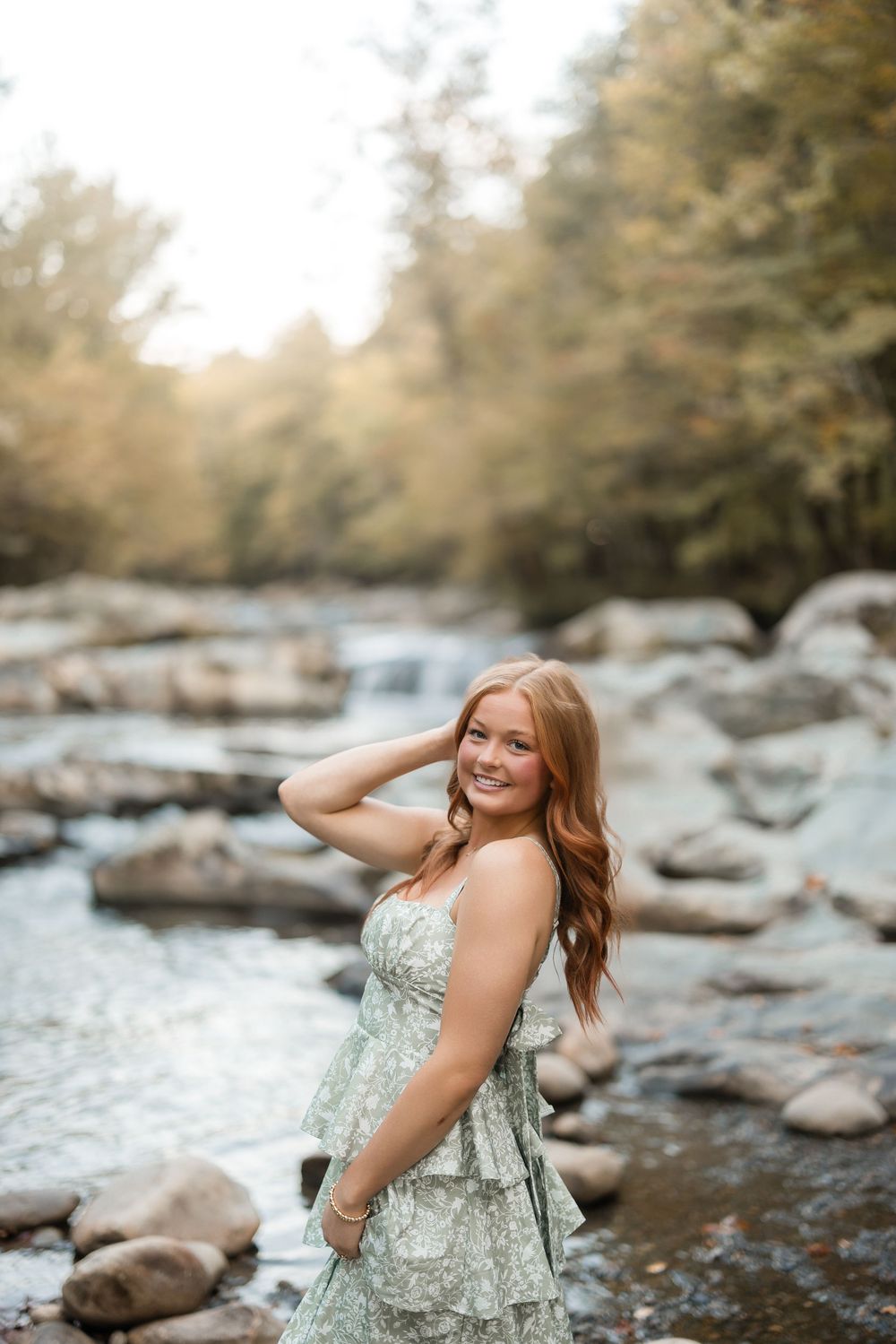 A woman in a floral dress poses playfully by a rocky riverbed surrounded by trees.