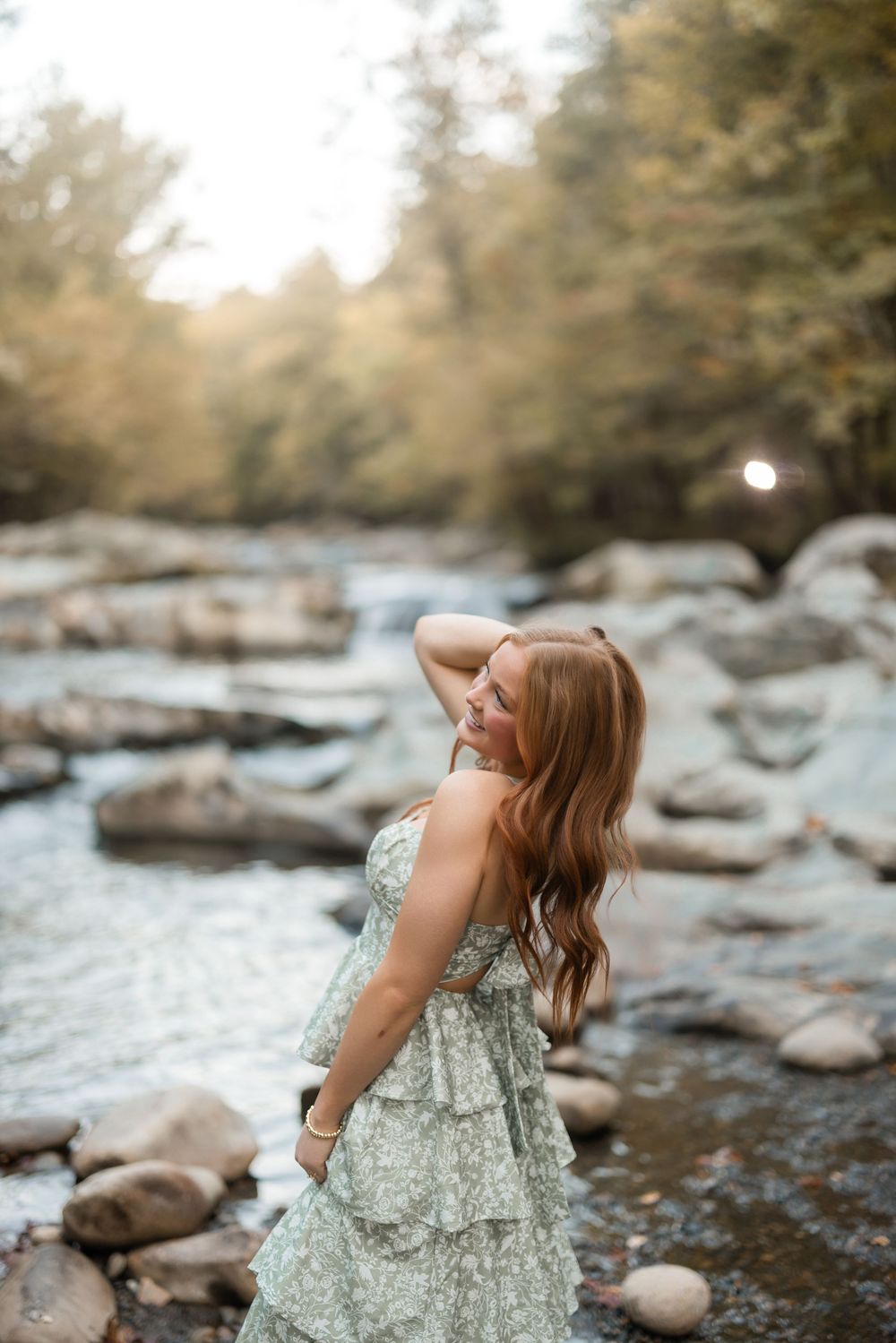 Person in a floral dress sits thoughtfully by a rocky riverbank surrounded by autumn trees and flowing water.