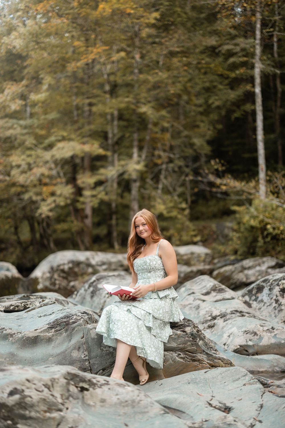 A person in a floral dress reclines on large rocky outcrops by a river surrounded by autumn forest scenery.