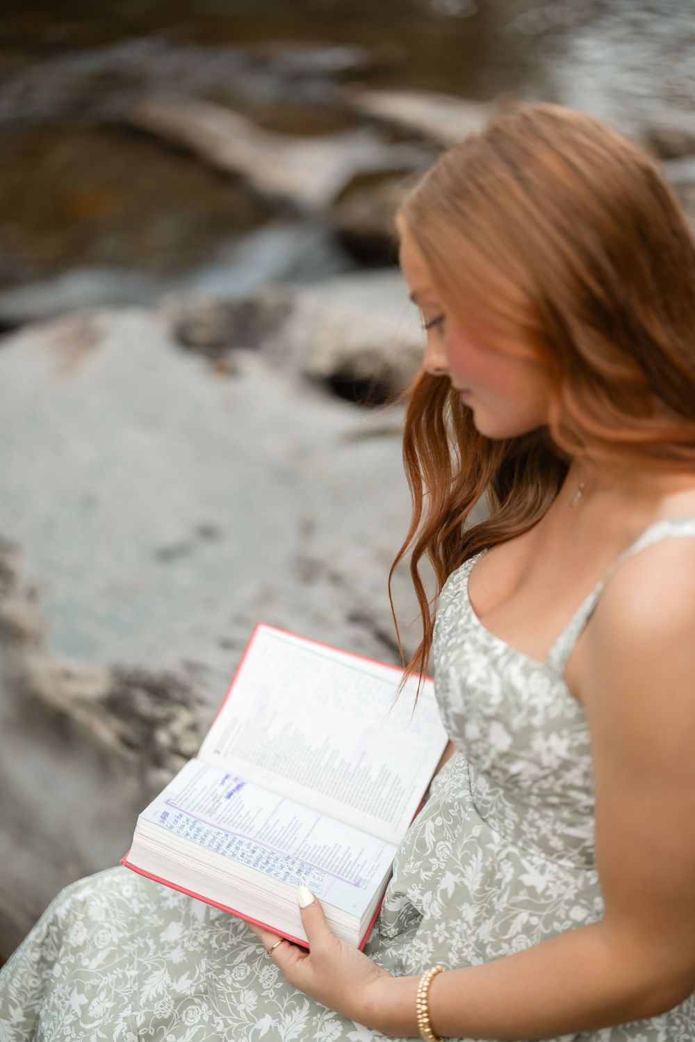 A person in a white dress holds an open book while sitting on a rocky surface near water.