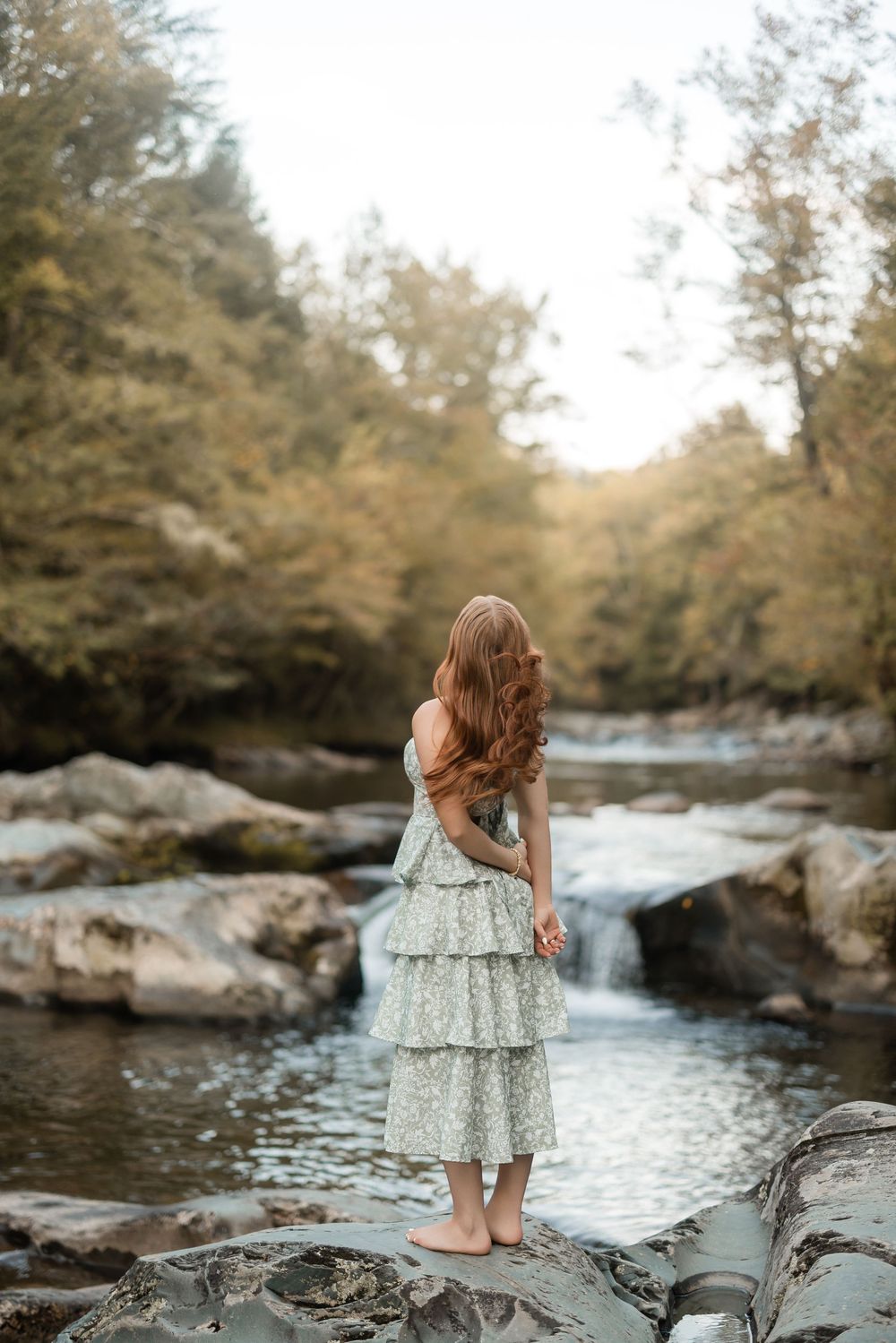 A person in a tiered blue dress stands on rocks by a flowing river in an autumn forest setting.