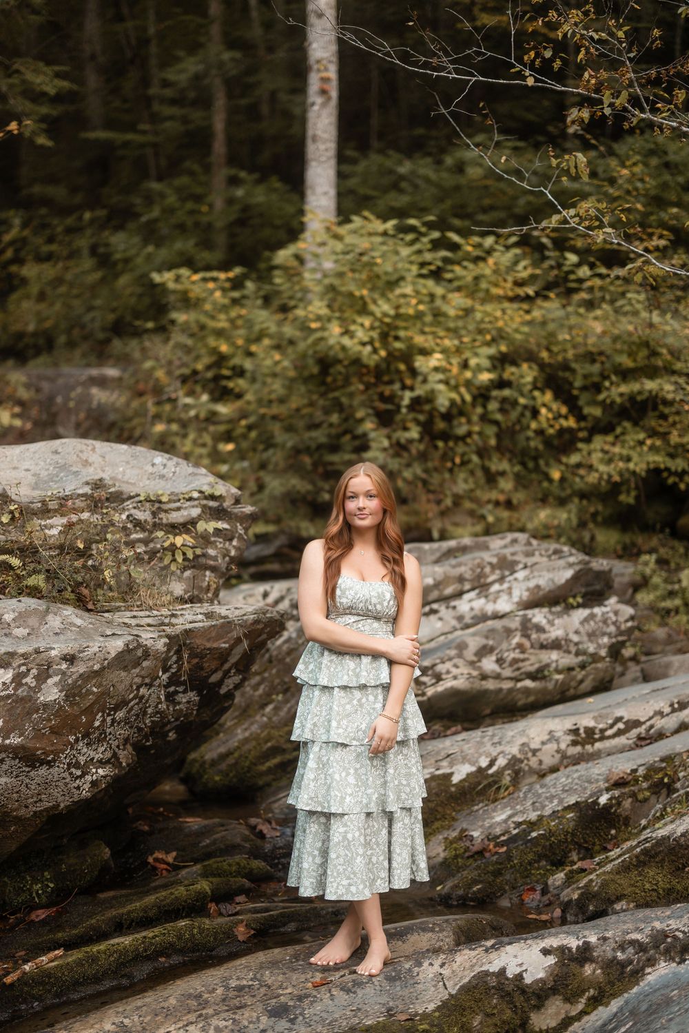 Barefoot pose on rocky riverbed wearing a tiered floral maxi dress in a forest setting.