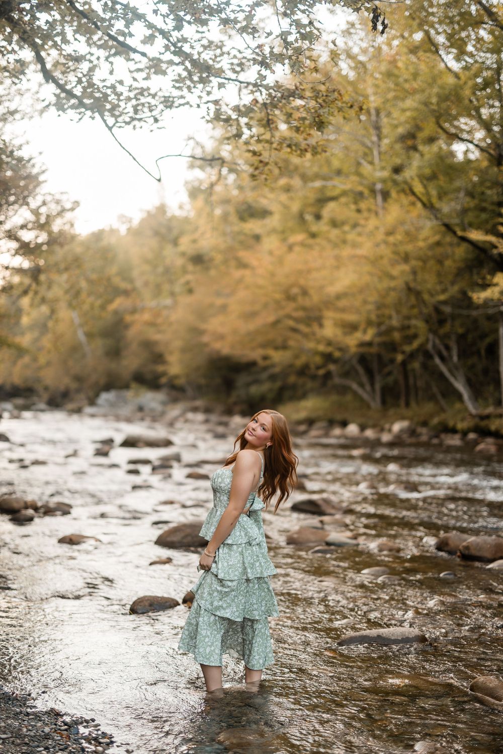 A person in a flowing mint dress stands barefoot in a rocky stream surrounded by autumn trees.
