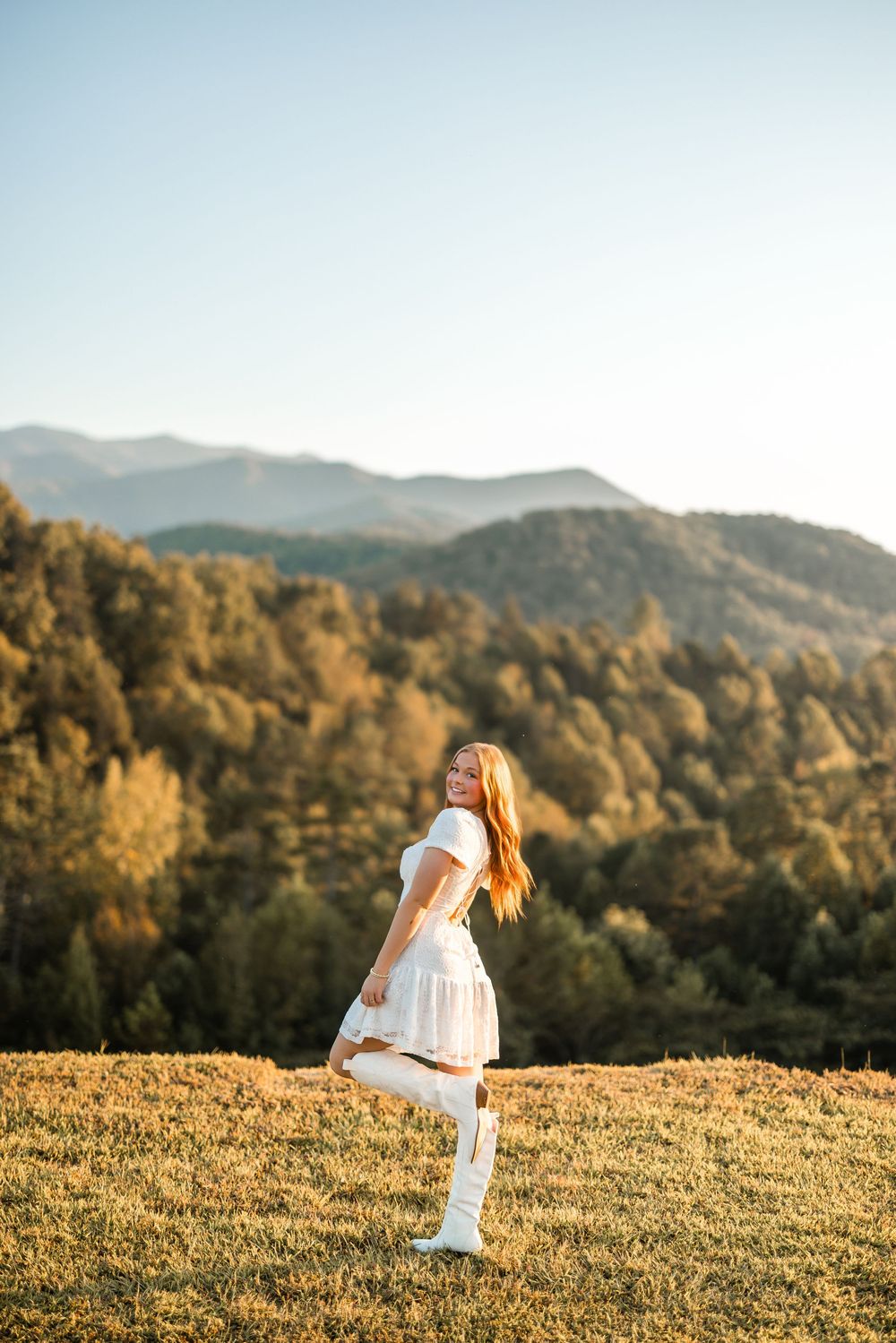 A figure in a white dress stands in a sunlit meadow with golden mountains and autumn trees in the background.