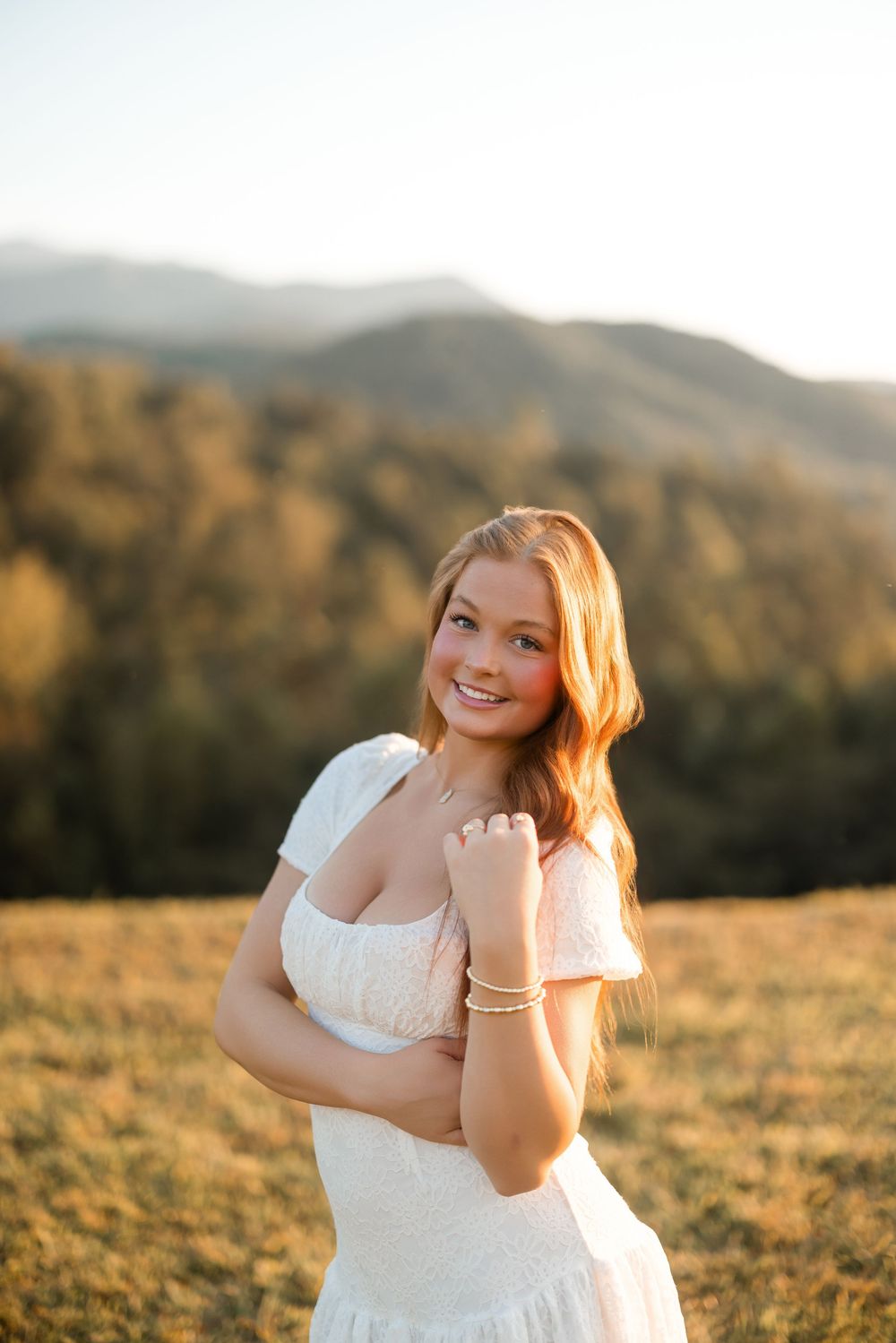 A person in a white dress poses in a golden field at sunset with mountains in the background.
