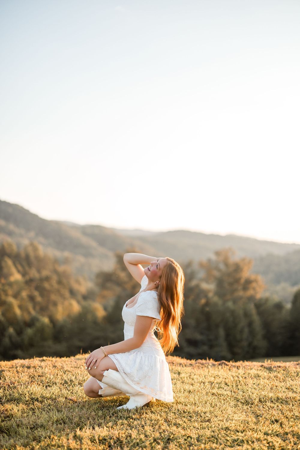 A person in a white dress sits on a grassy hillside during golden hour with mountains in the background.