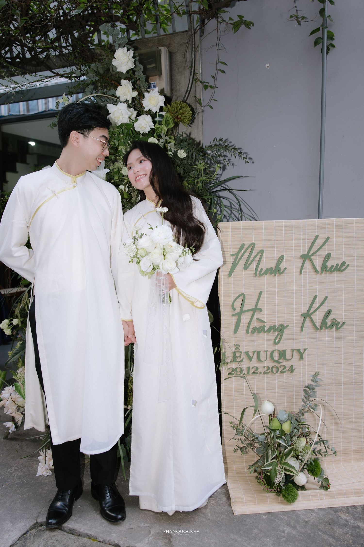 Vietnamese bride and groom in white ao dai share a loving glance at their Le Vu Quy ceremony.