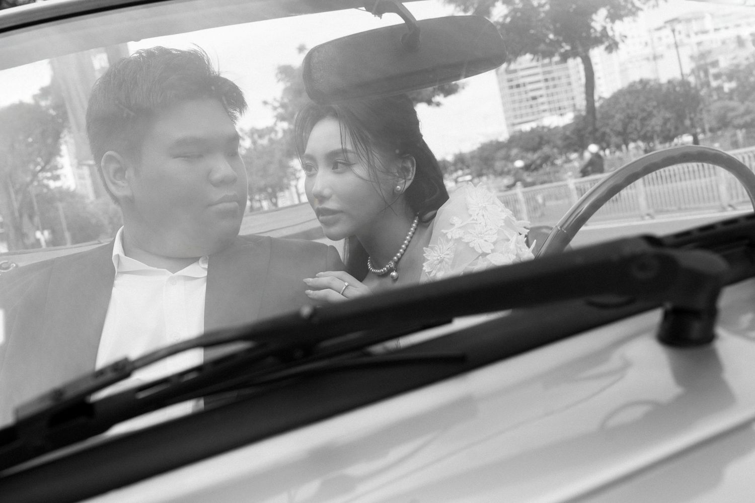 Black and white photo of a bride and groom sitting together inside a car, captured through the window.