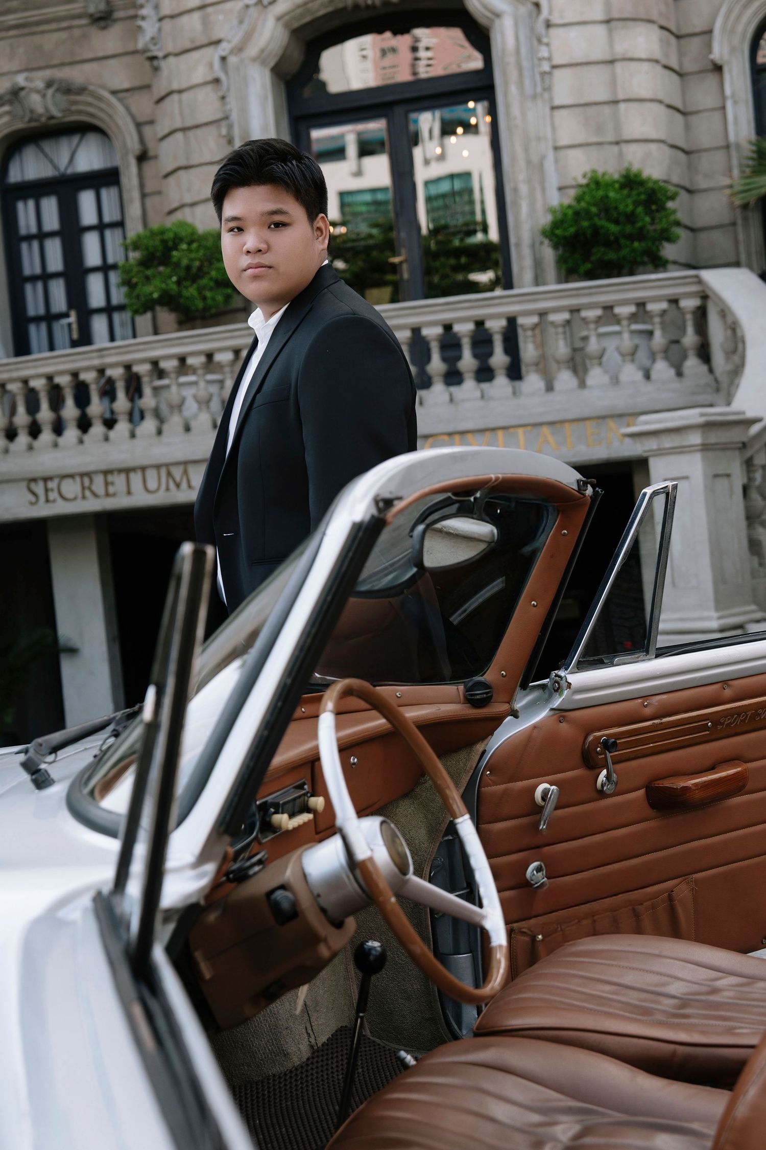 Young Asian man in black suit poses beside a vintage white convertible car outside an elegant building.
